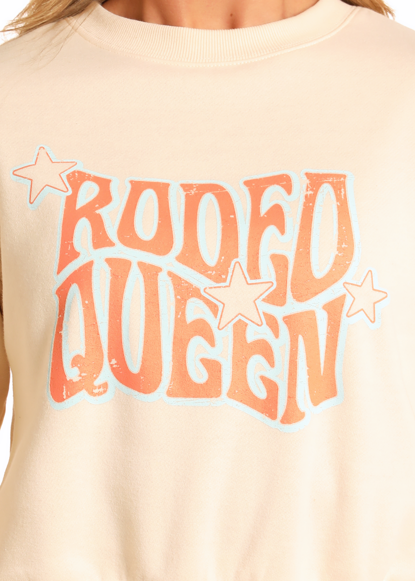 A person wears the Rock & Roll Denim Rodeo Queen Graphic Pullover—cream-colored with bold retro orange text, blue outline, and three cream stars—a relaxed-fit statement style.