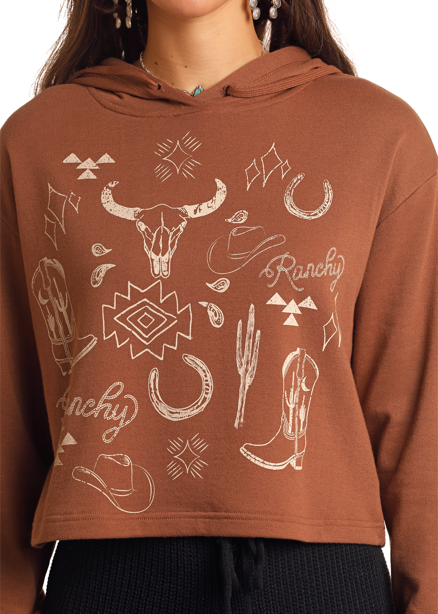 A person wears the Rock & Roll Denim Range Western Graphic Cropped Hoodie in brown, with white bull skull, cowboy boots, hats, cacti, and horseshoes graphics plus "Ranchy" text. Only the torso and part of the face are shown.