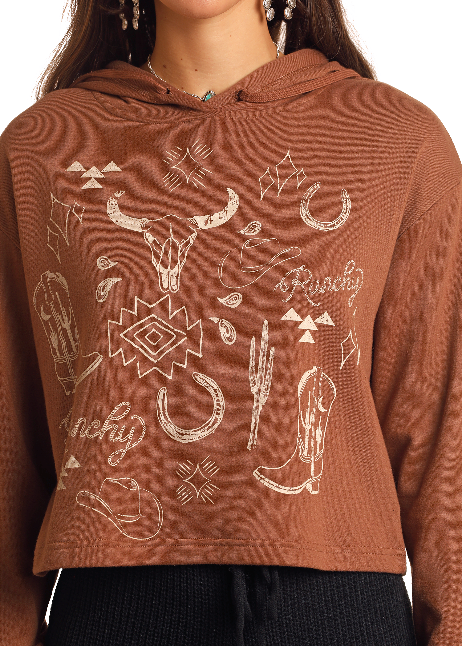 A person wears the Rock & Roll Denim Range Western Graphic Cropped Hoodie in brown, with white bull skull, cowboy boots, hats, cacti, and horseshoes graphics plus "Ranchy" text. Only the torso and part of the face are shown.