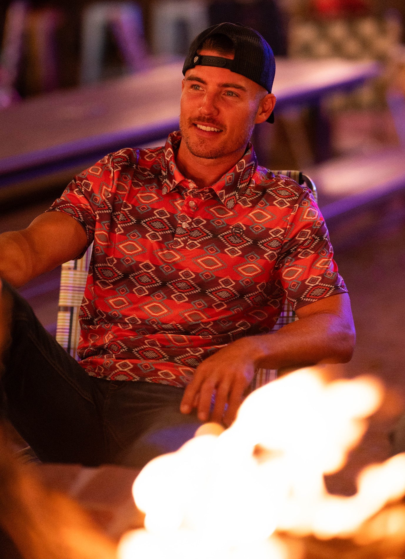 A man in a Rock & Roll Denim Aztec Vertical Striped Red Polo (S) and a backwards cap sits relaxed by a campfire, smiling in a warmly lit outdoor setting.