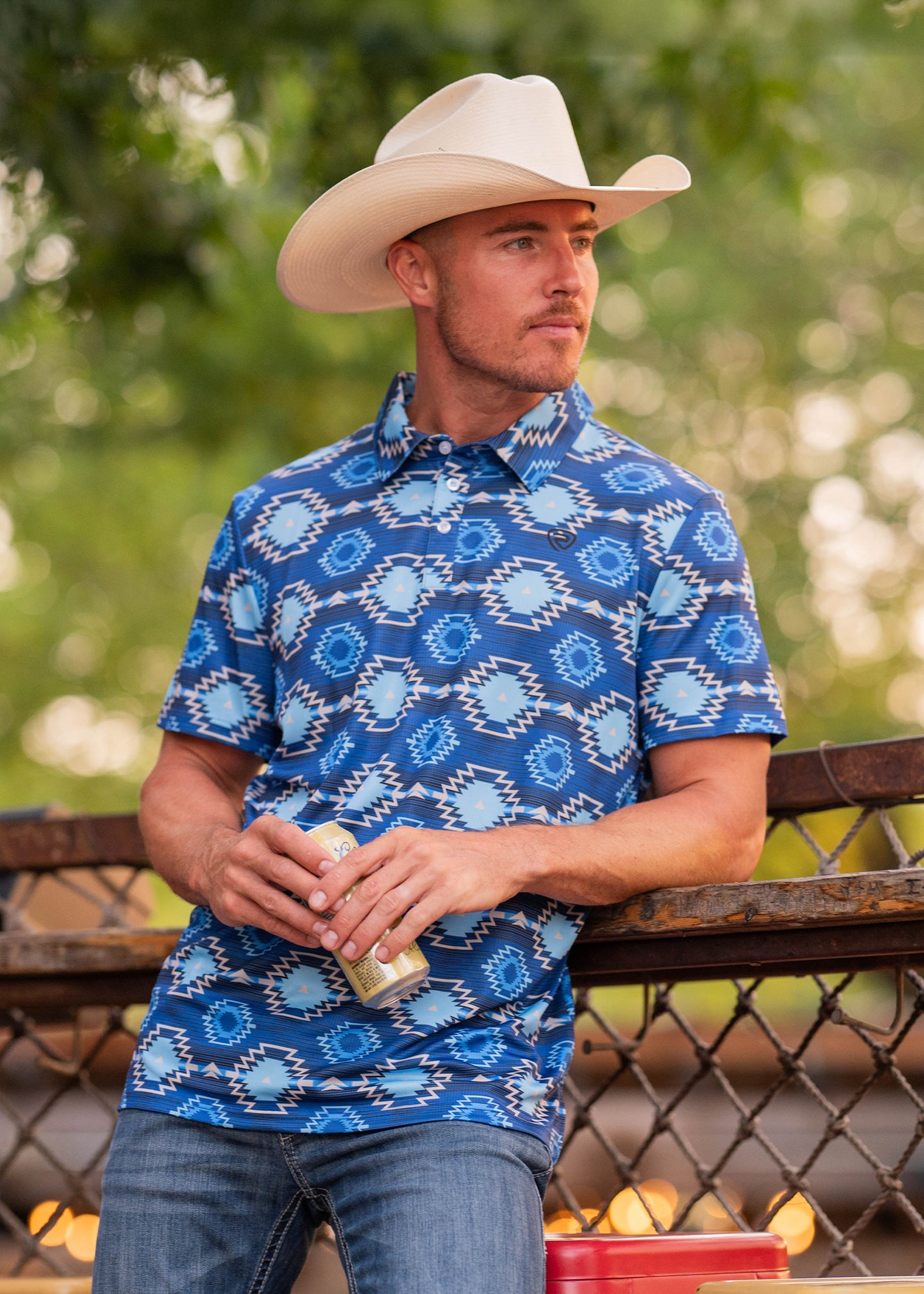 A man in a Rock & Roll Denim Bold Blue Aztec Polo and a white cowboy hat leans on a wooden fence, holding a canned drink. Trees and a blurred background create an outdoor, relaxed vibe.