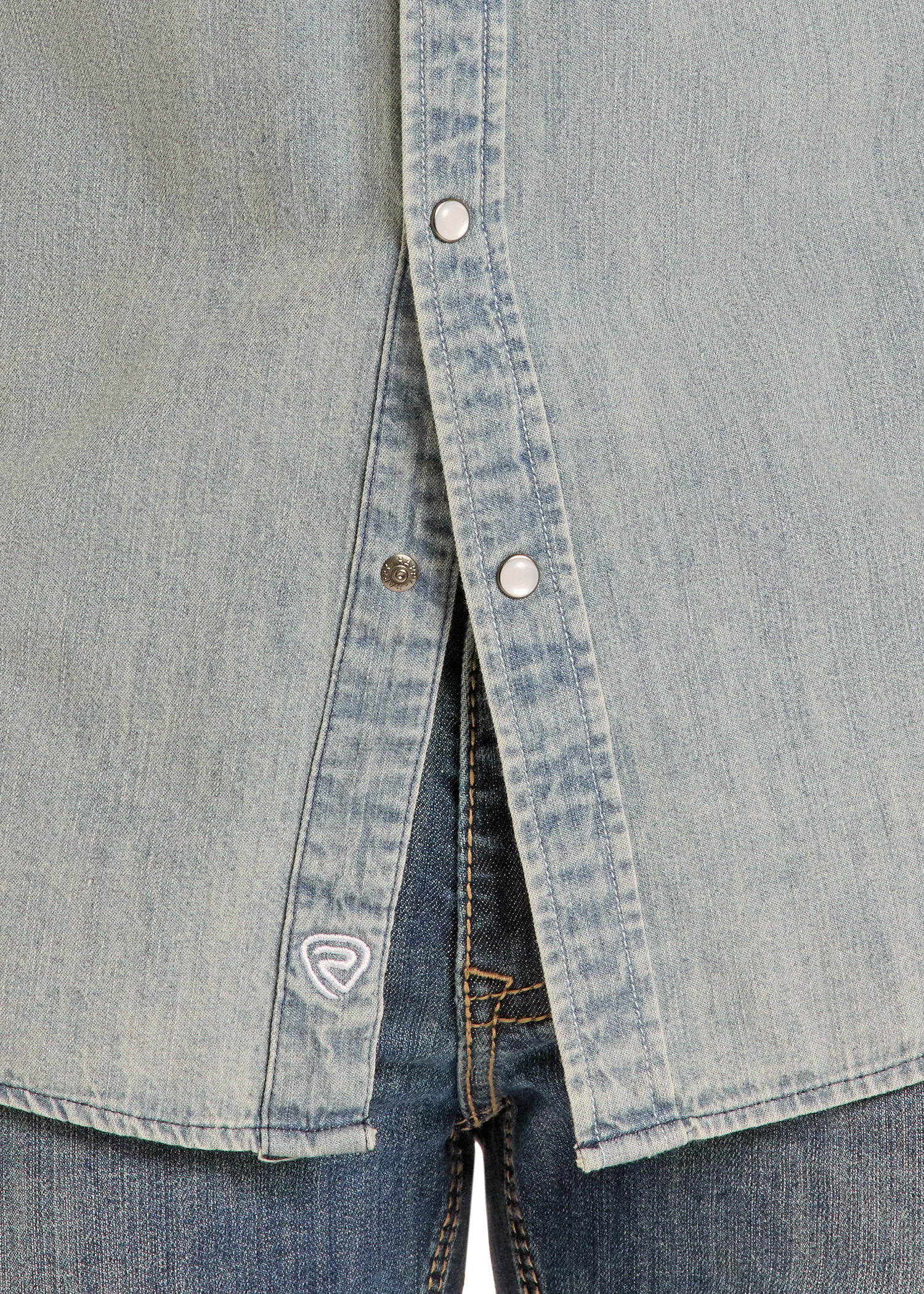 Close-up of the lower part of the Rock & Roll Denim Slim Fit Medium Wash Denim Snap Shirt with silver snap buttons, worn over blue jeans. A small embroidered logo is visible near the hem.