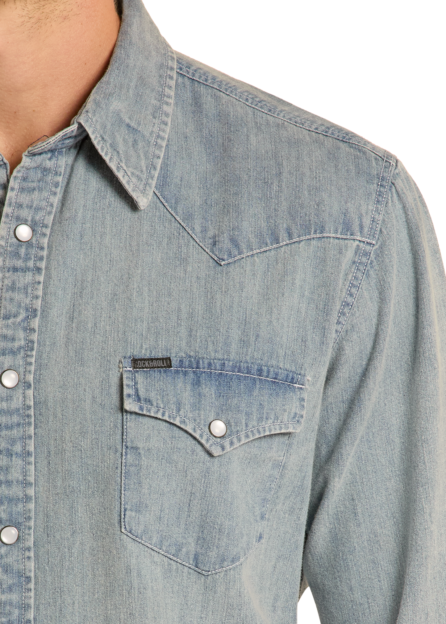 A close-up of the Rock & Roll Denim Slim Fit Medium Wash Denim Snap Shirt, featuring silver snap buttons, a pointed collar, buttoned chest pocket, and western-style stitching, with part of the wearer’s neck and shoulder visible.