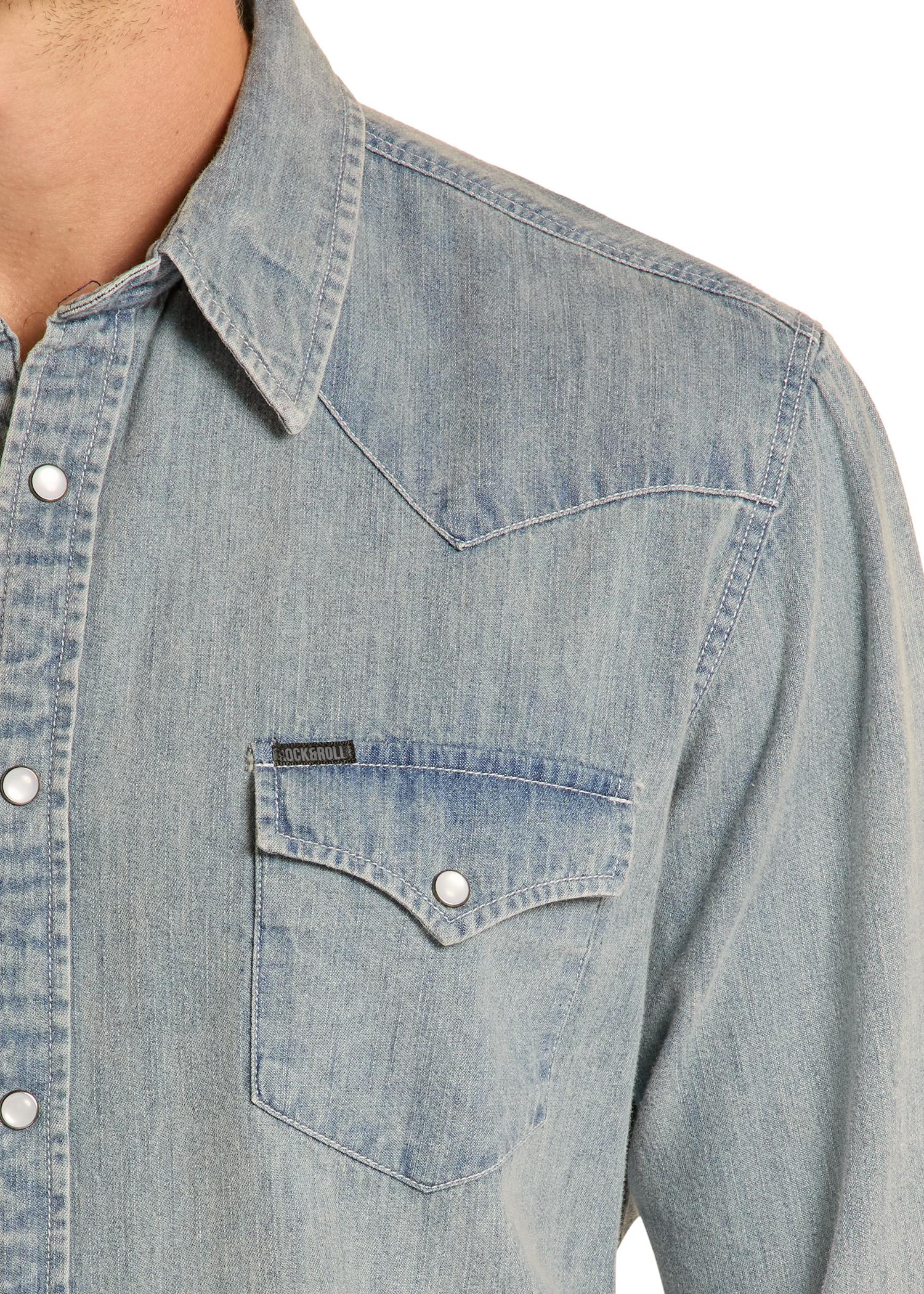 A close-up of the Rock & Roll Denim Slim Fit Medium Wash Denim Snap Shirt, featuring silver snap buttons, a pointed collar, buttoned chest pocket, and western-style stitching, with part of the wearer’s neck and shoulder visible.