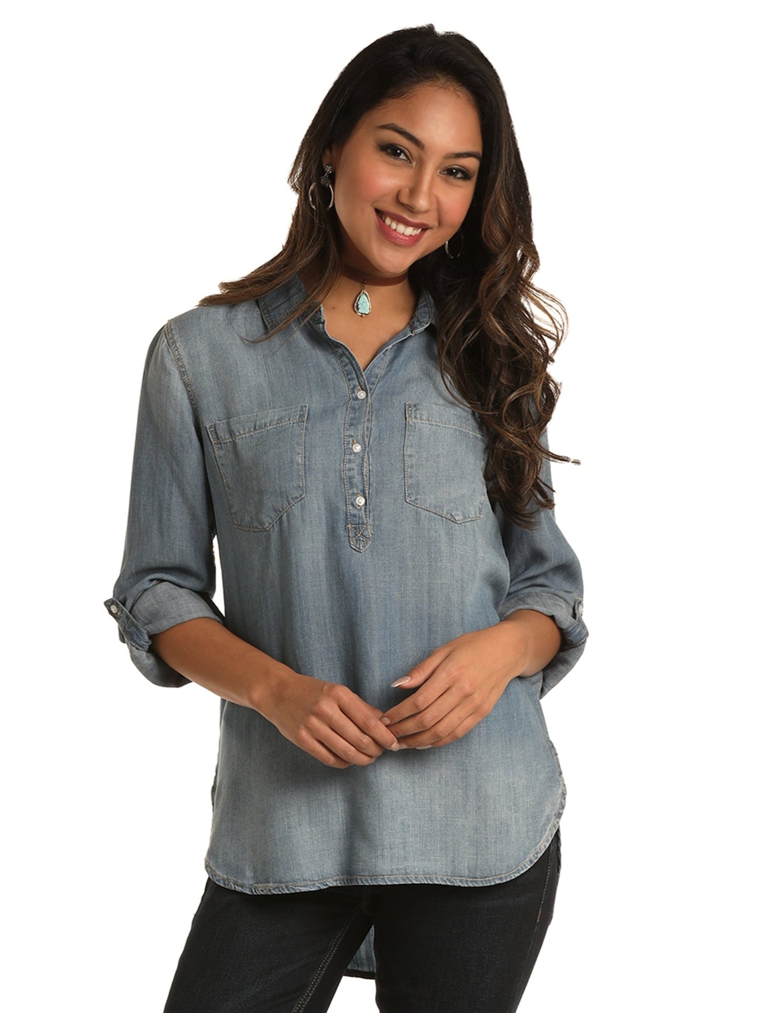 A woman with long wavy hair smiles while wearing the Rock & Roll Denim Denim Blouse in a relaxed fit light blue wash with rolled sleeves and black pants, standing against a white background.