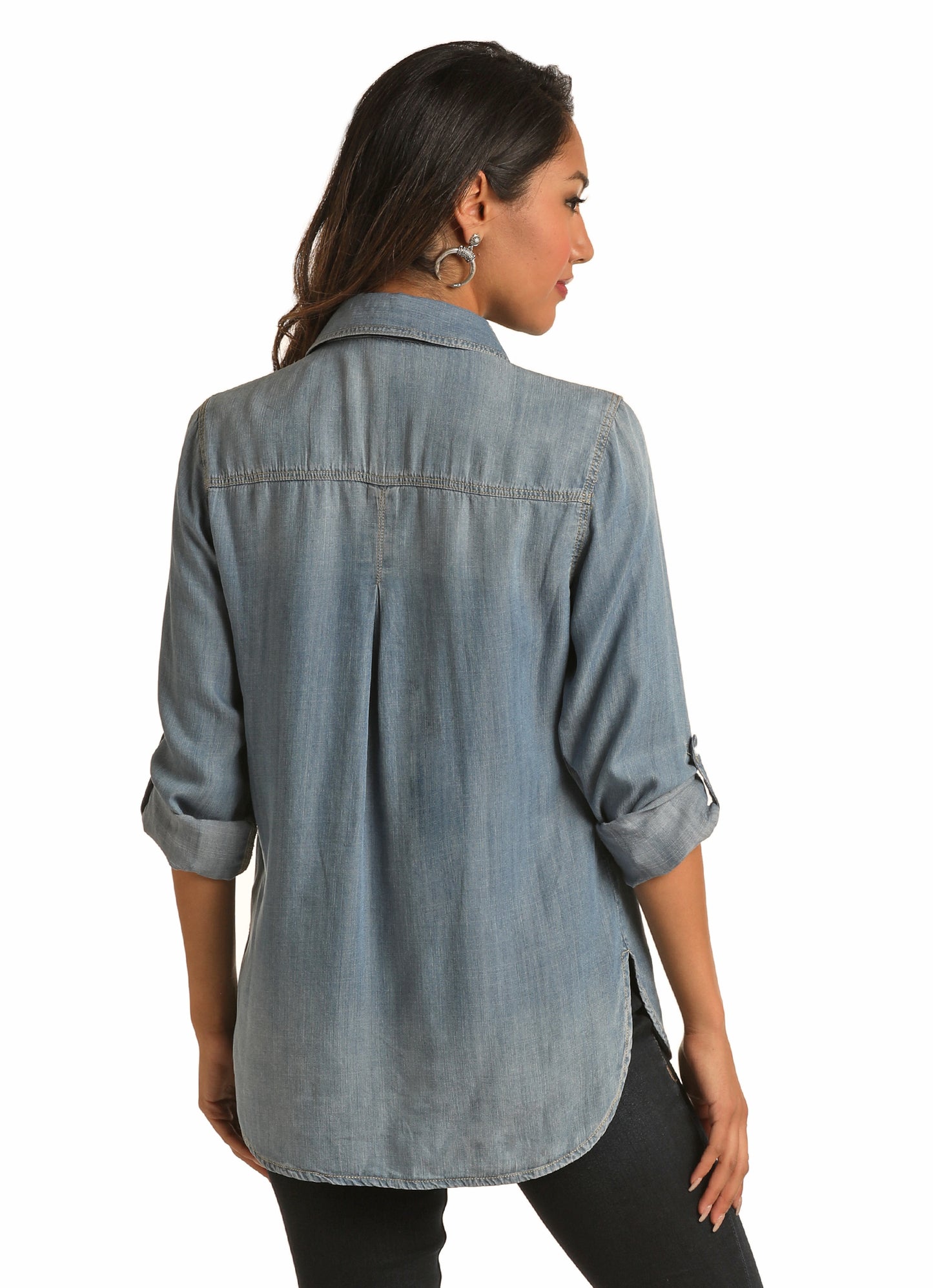 A woman with long dark hair stands with her back to the camera, wearing the Rock & Roll Denim Denim Blouse in light blue with rolled-up sleeves and a relaxed fit, paired with black pants against a white background.