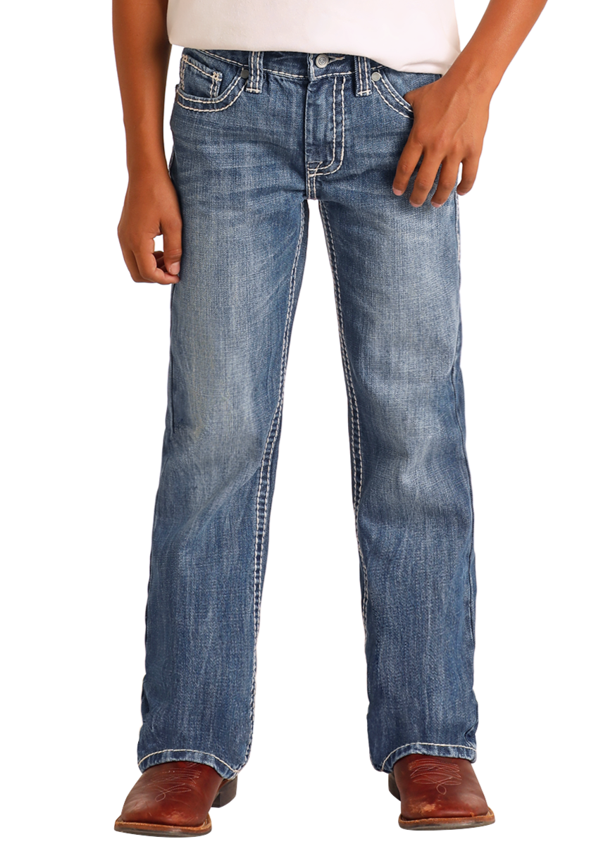 Wearing Rock & Roll Denim Boys' Ivory Top-Stitching Regular Fit Bootcut Jeans, a white shirt, and brown cowboy boots, a person stands against a plain background.