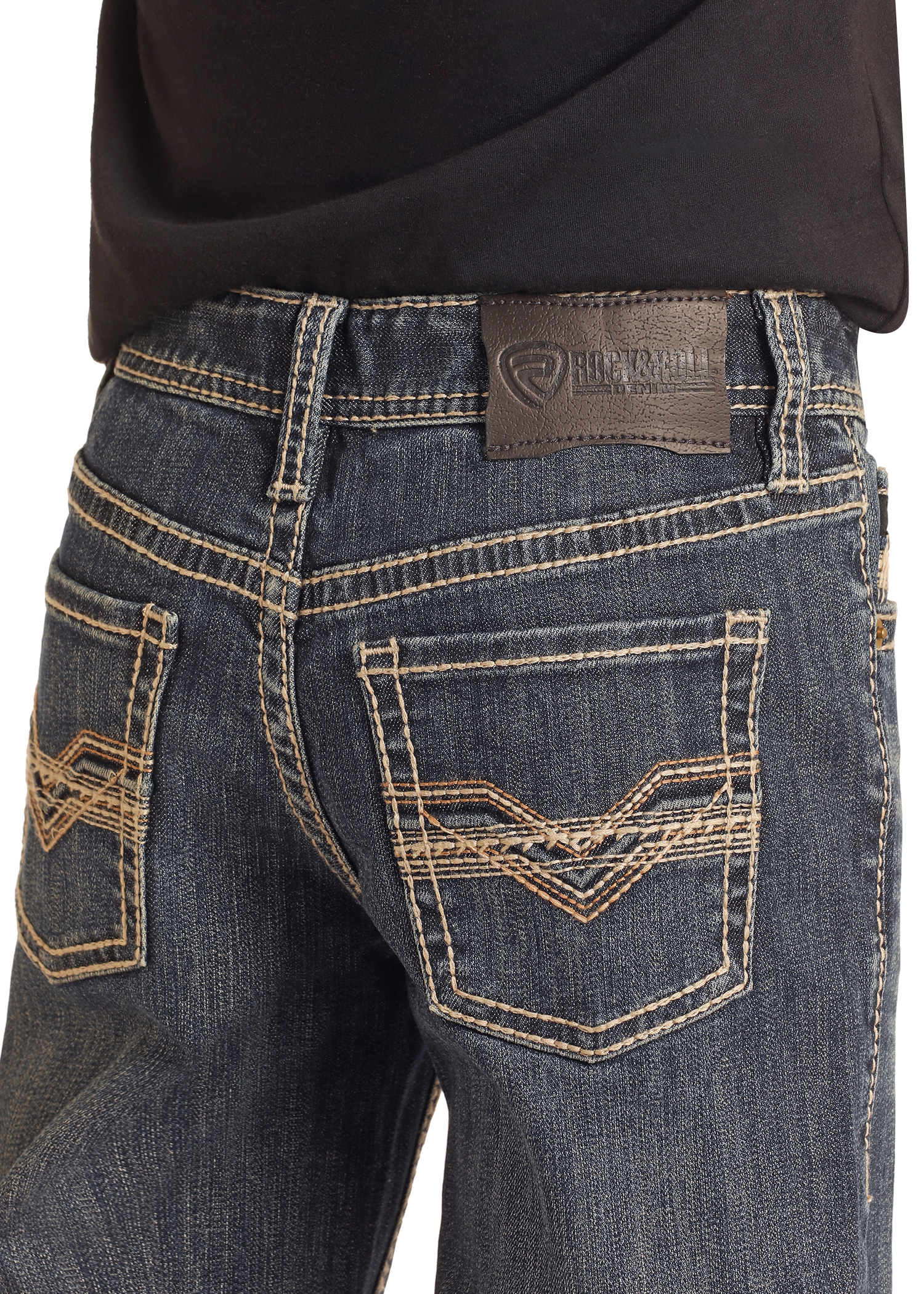 Close-up of a person wearing Rock & Roll Denim Boys' Regular Fit Raised Embroidery Bootcut Jeans in dark blue, featuring tan stitching, decorative pocket designs, and a black leather patch on the waistband under a black shirt.