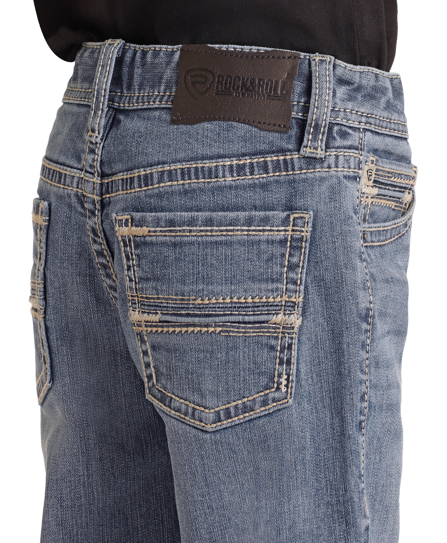 A close-up shows a person in a black shirt tucked into Boys' Regular Fit Two-Tone Embroidered Bootcut Jeans by Rock & Roll Denim, featuring detailed stitching and a black brand patch above the right pocket.