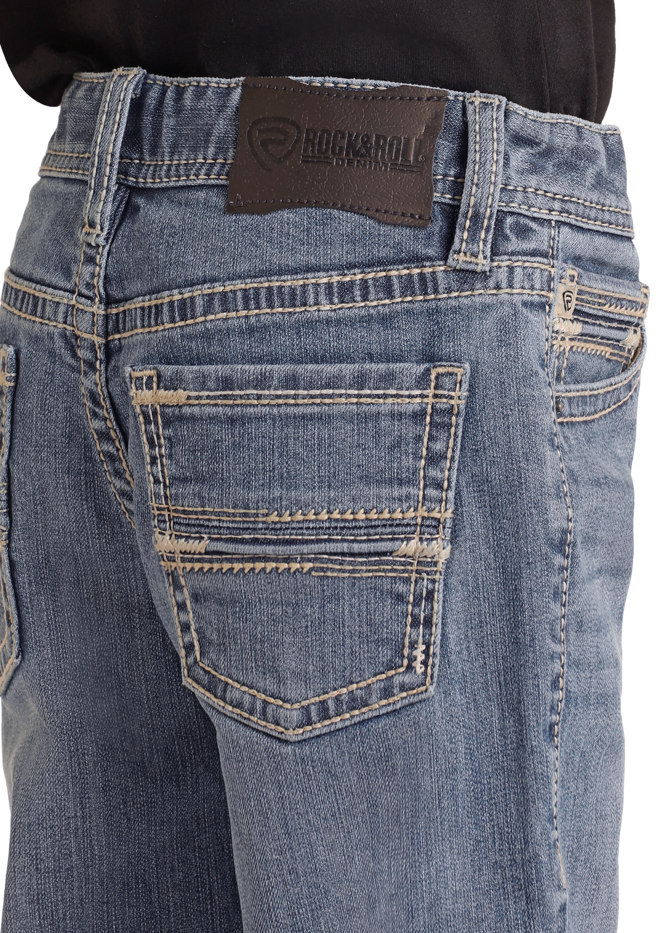 A close-up shows a person in a black shirt tucked into Boys' Regular Fit Two-Tone Embroidered Bootcut Jeans by Rock & Roll Denim, featuring detailed stitching and a black brand patch above the right pocket.