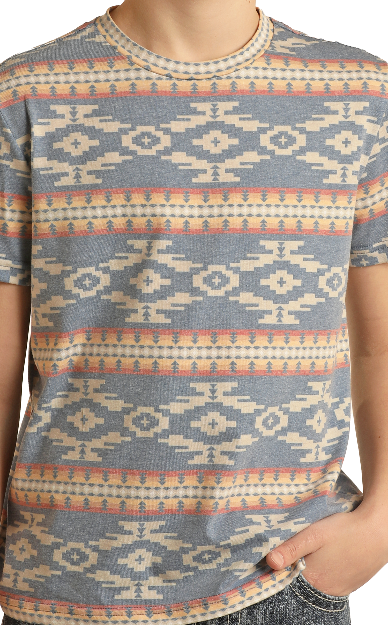 A person wears a Rock & Roll Denim Boys' Aztec Print T-Shirt featuring a blue, beige, and orange geometric southwestern pattern. One hand rests in their jeans pocket and only their upper body is shown.