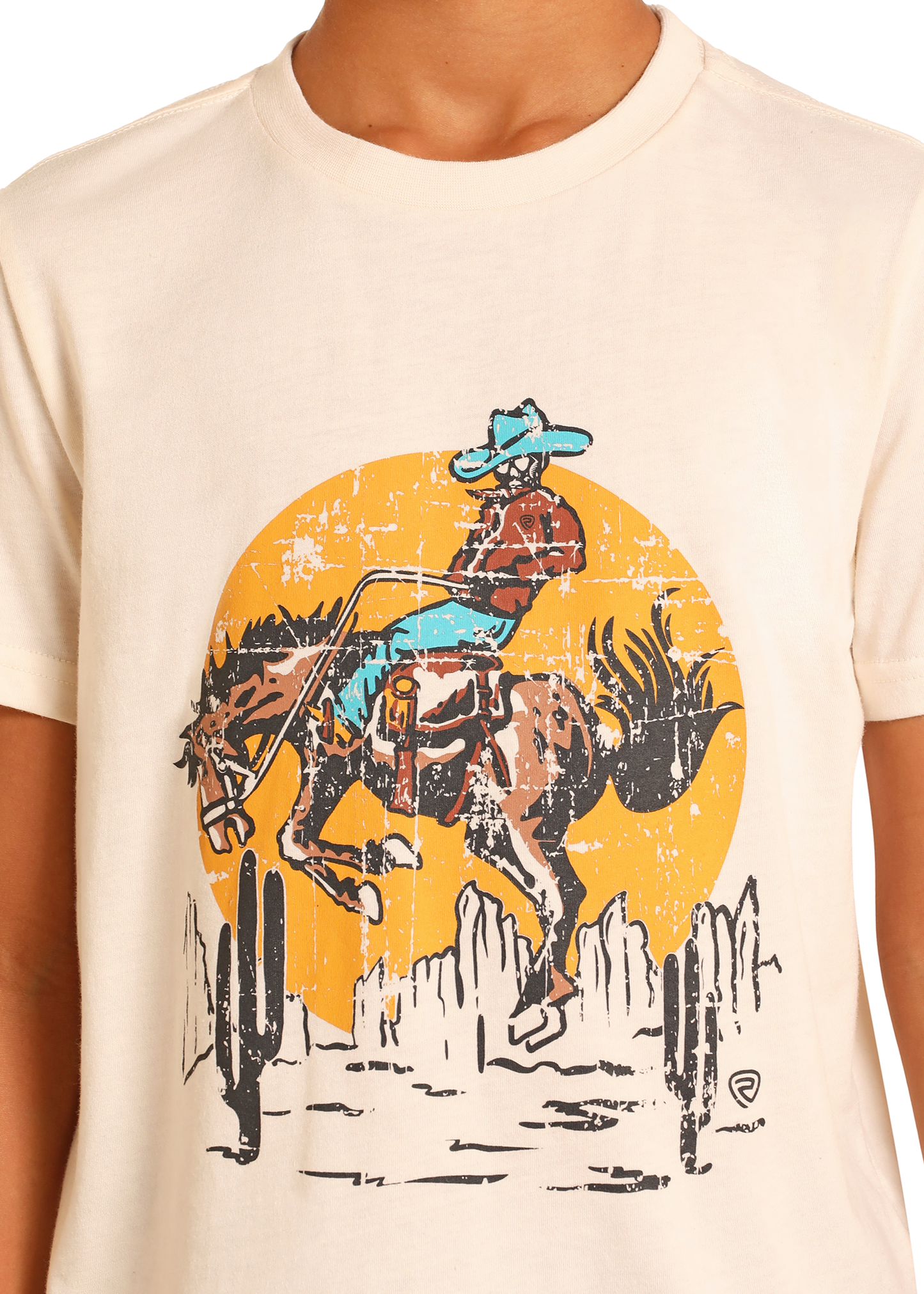 A boy wears the Rock & Roll Denim Boys' Buckin' Bronc Graphic Tee, featuring a cowboy riding a bucking horse with cacti and a yellow sun in the background.