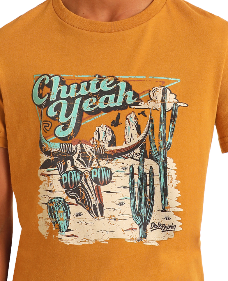 A person wearing the Rock & Roll Denim Dale Brisby Desert Graphic Tee, a men's cotton T-shirt with a desert skull, cacti, cow skull graphic, and "Chute Yeah" plus "Pow Pow" boldly on the front.