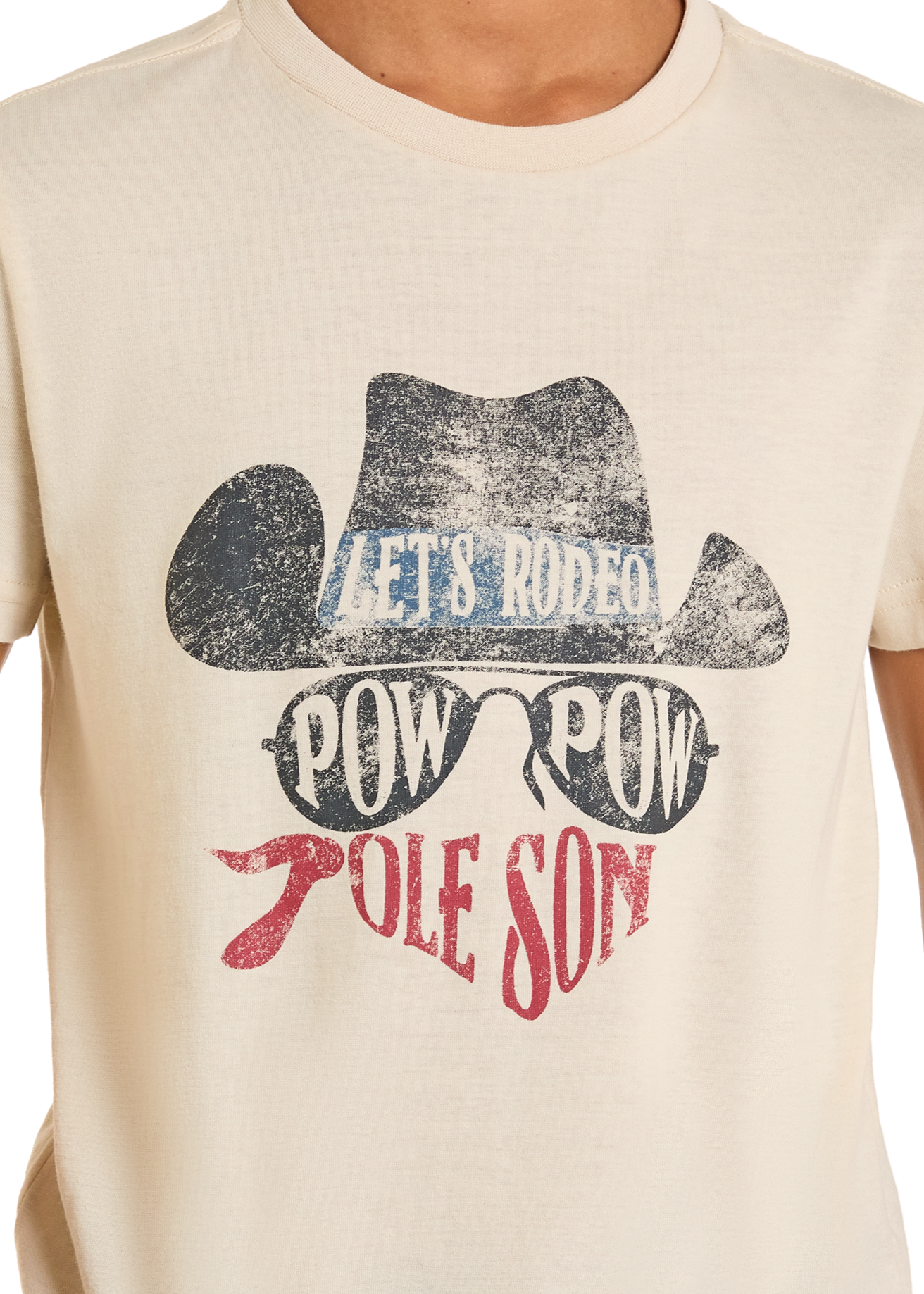 A person wears the Boys' Dale Brisby Pow Pow Graphic Tee by Rock & Roll Denim, a cream shirt with a cowboy hat, sunglasses, and red bandana graphic plus "LET’S RODEO," "POW POW," and "OLE SON" in bold colors.