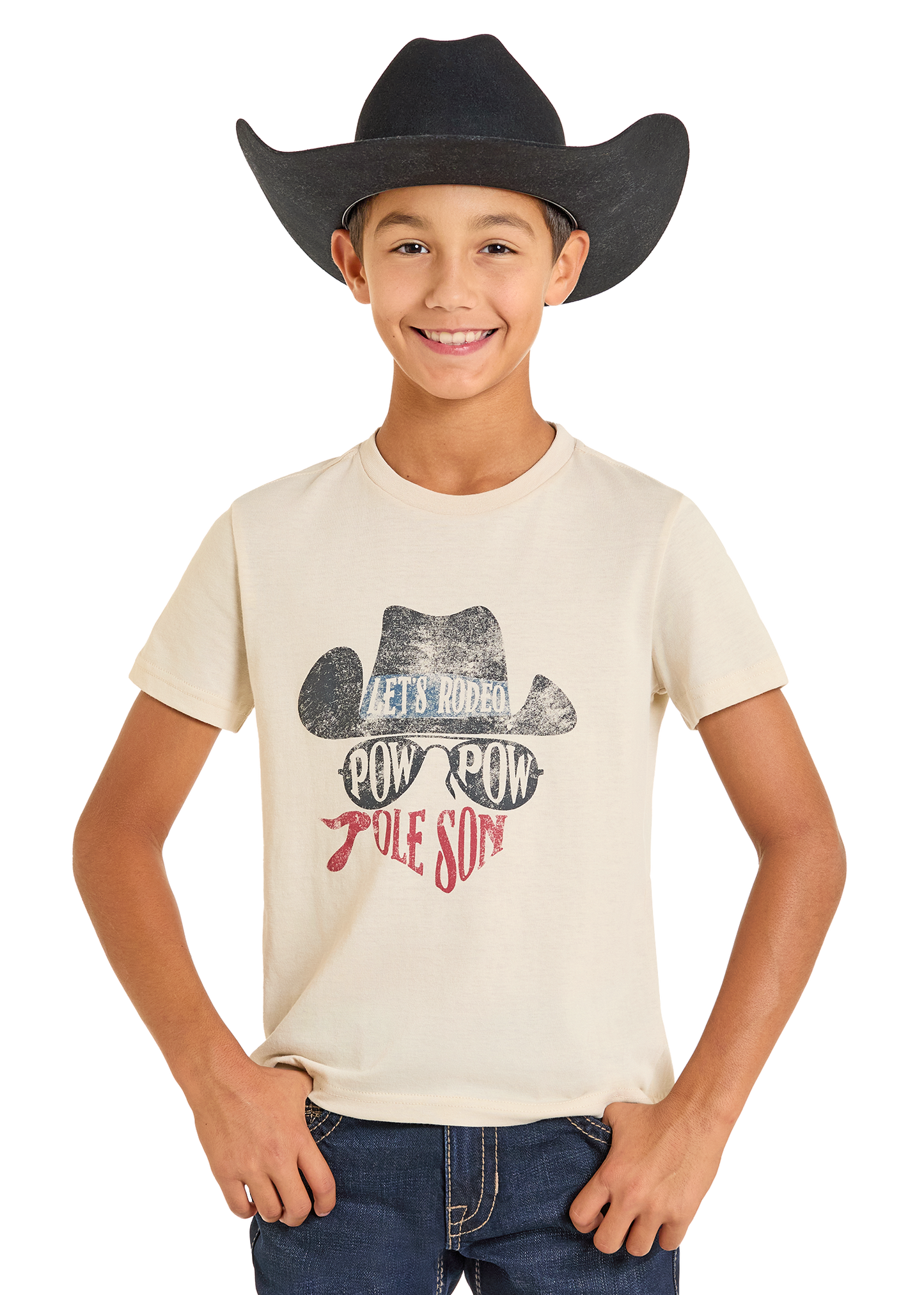 A smiling boy wears a black cowboy hat and a cream Rock & Roll Denim Boys' Dale Brisby Pow Pow Graphic Tee featuring a cowboy hat graphic and "LETS RODEO POW POW POLE SON" text, with his hands tucked in his jean pockets.