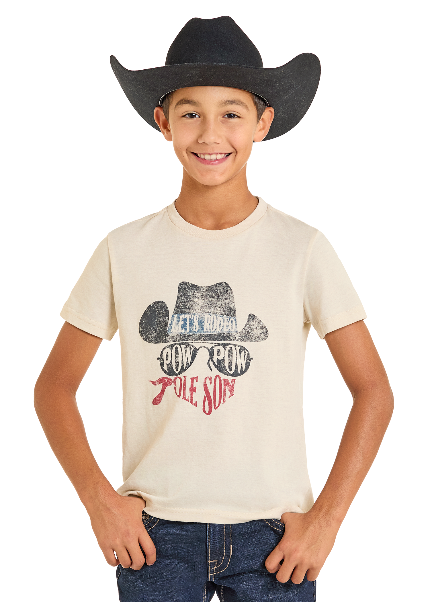 A smiling boy wears a black cowboy hat and a cream Rock & Roll Denim Boys' Dale Brisby Pow Pow Graphic Tee featuring a cowboy hat graphic and "LETS RODEO POW POW POLE SON" text, with his hands tucked in his jean pockets.