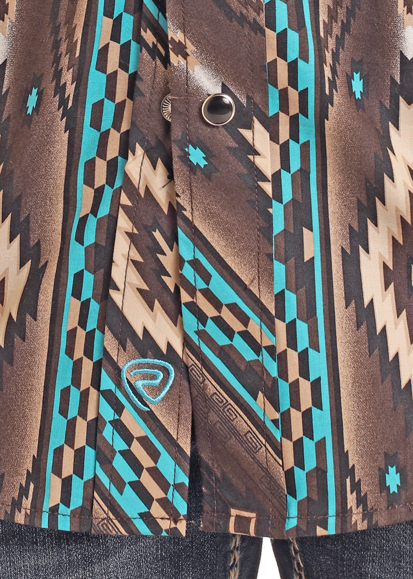 A close-up of the Rock & Roll Denim Boys' Southwest Print Long Sleeve Snap Shirt shows its geometric brown, tan, and turquoise pattern with snap buttons and an embroidered turquoise logo above blue denim jeans.