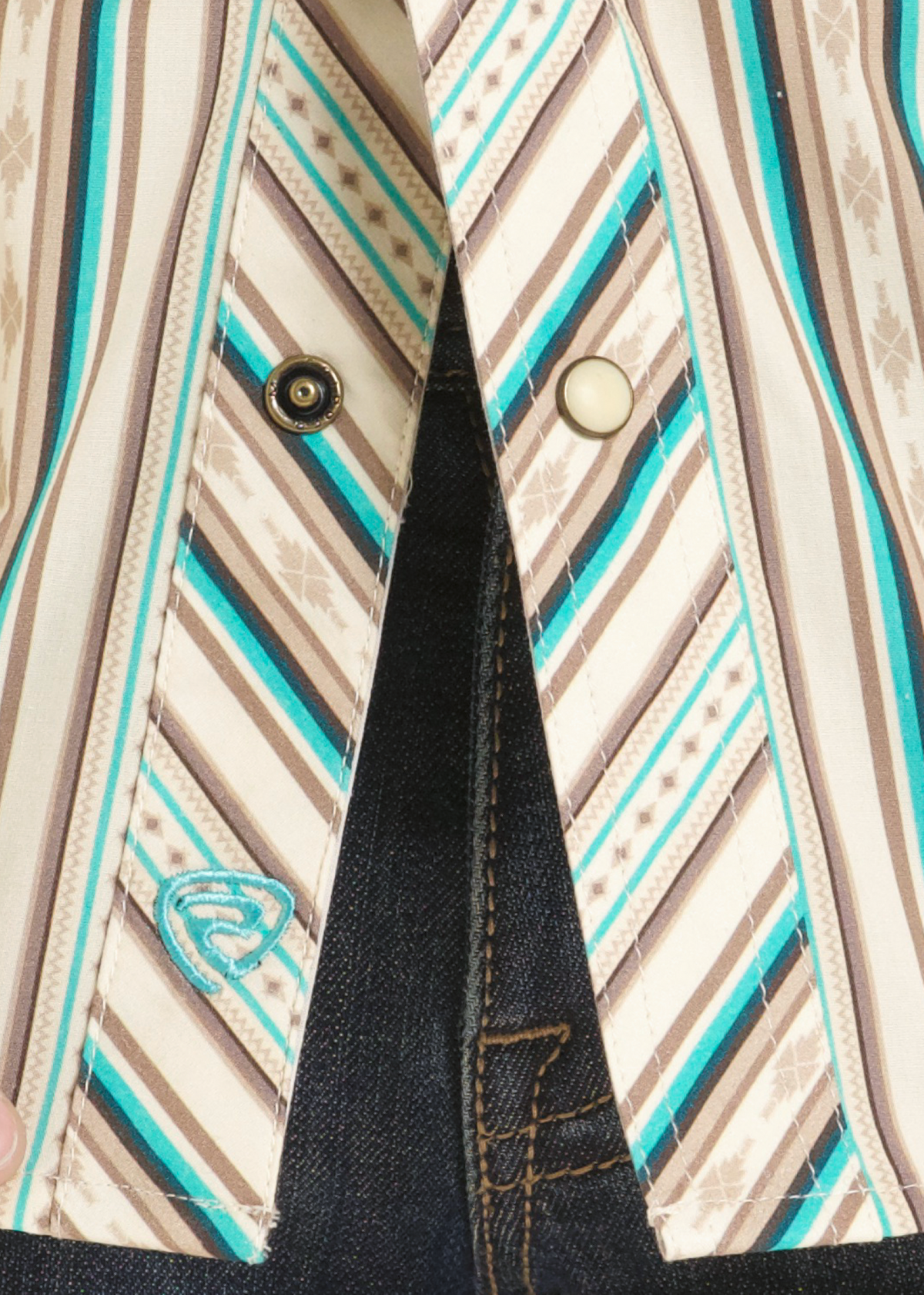 Close-up of a striped button-up shirt with diagonal bands of teal, brown, beige, and white, paired with dark blue jeans. A small embroidered logo is visible on the shirt pocket.