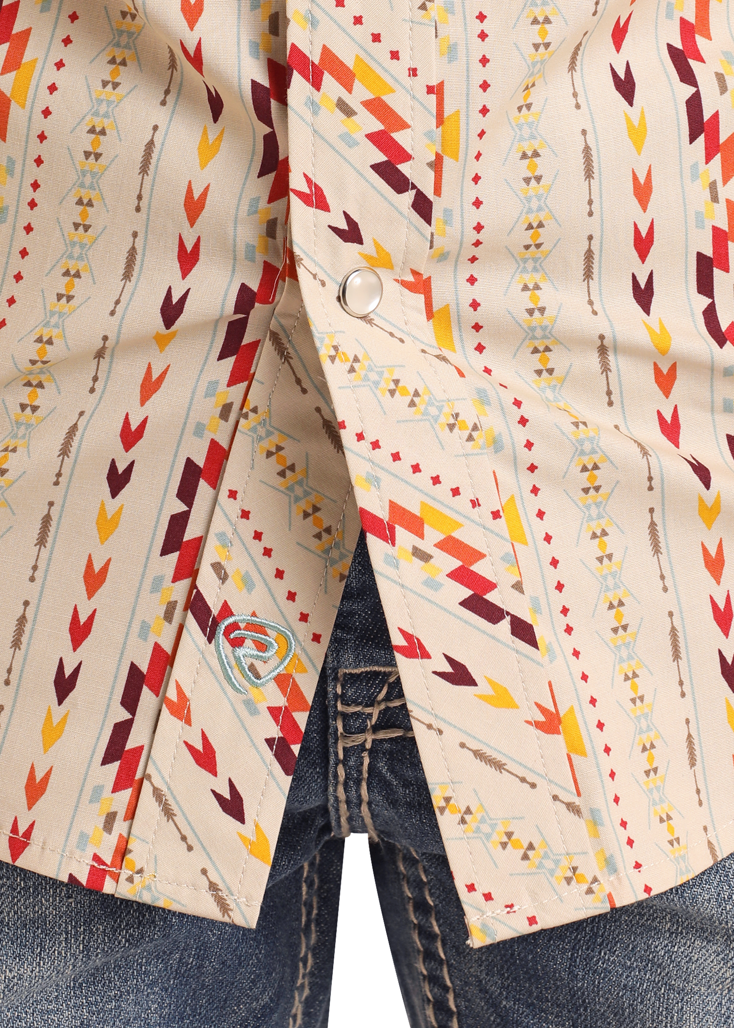 A close-up of the Rock & Roll Denim Boys' Vertical Aztec Short Sleeve Snap Shirt, featuring a vibrant red, yellow, and brown geometric pattern and arrow designs, partially snapped over denim jeans with a small logo patch at the hem.