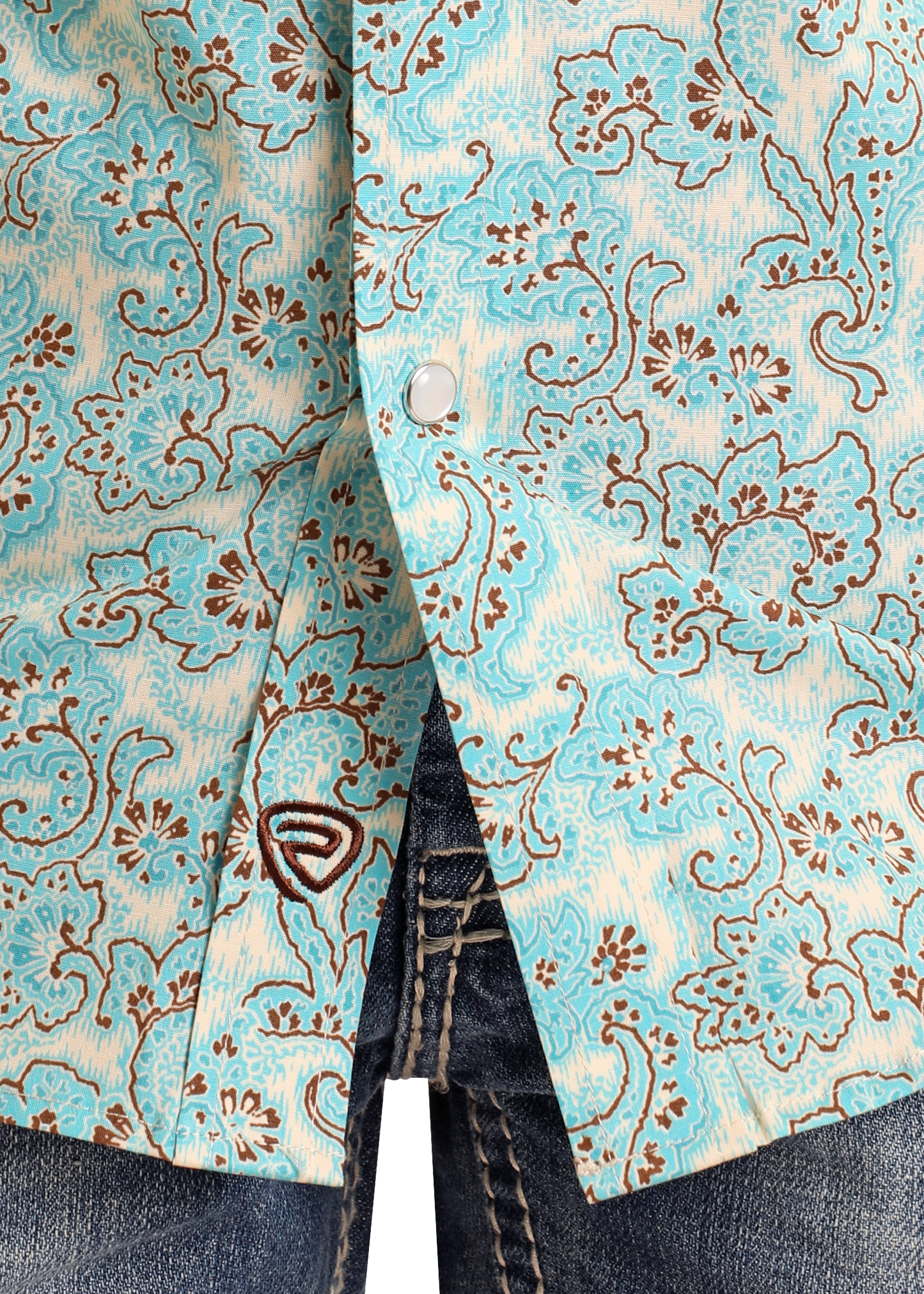 Close-up of a Rock & Roll Denim Boys' Paisley Short Sleeve Snap Shirt in turquoise and beige floral print over dark blue jeans. This Western shirt features snap buttons, a brown embroidered logo, and intricate paisley patterns on cotton spandex.