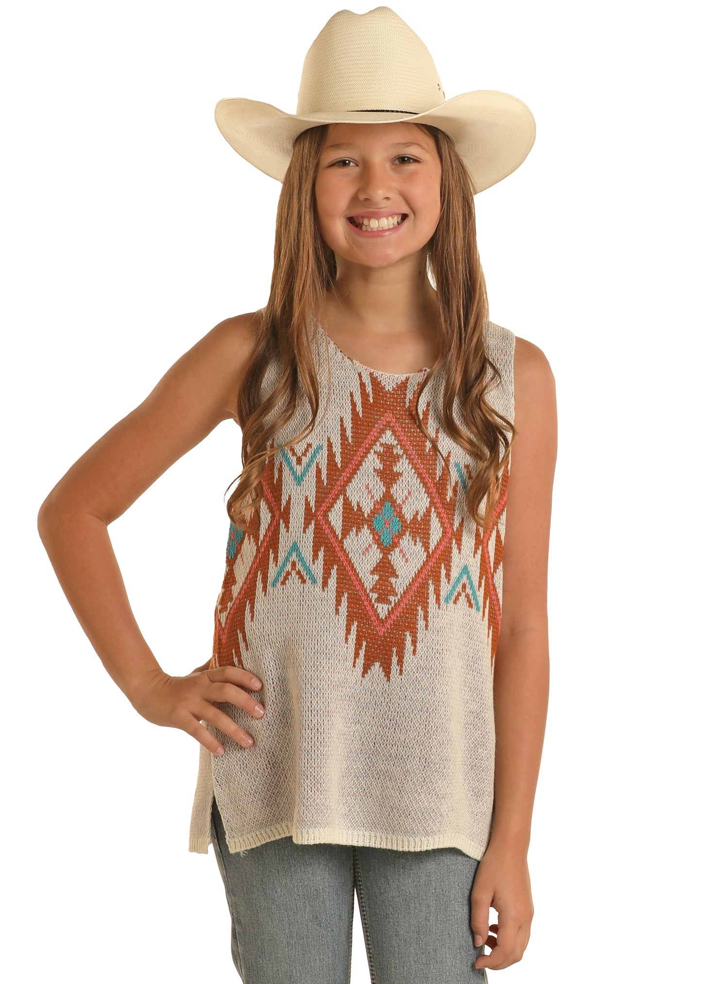 A smiling girl stands with one hand on her hip against a white background, wearing a white cowboy hat, blue jeans, and the Rock & Roll Denim Girls' Aztec Knit Tank featuring a colorful geometric pattern.