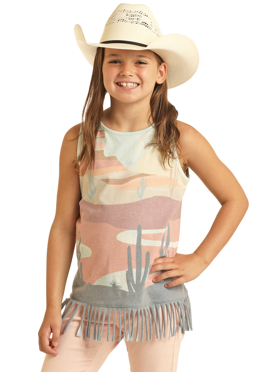 A smiling young girl wears the Rock & Roll Denim Girls' Desert Scenery Fringe Tank, featuring a desert print and fringe hem, paired with light pink pants and a white cowboy hat. She stands confidently with one hand on her hip against a plain background.