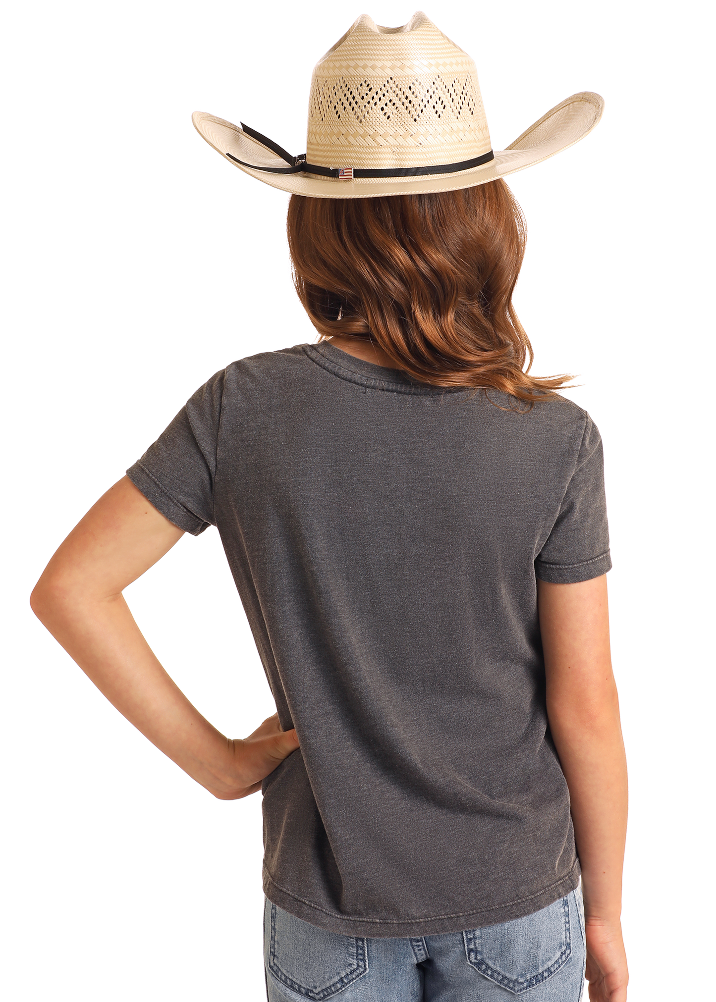 A person with wavy brown hair in a beige cowboy hat, blue jeans, and the Rock & Roll Denim Girls' Aztec Horse Graphic T-Shirt stands facing away with one hand on their hip against a plain background.