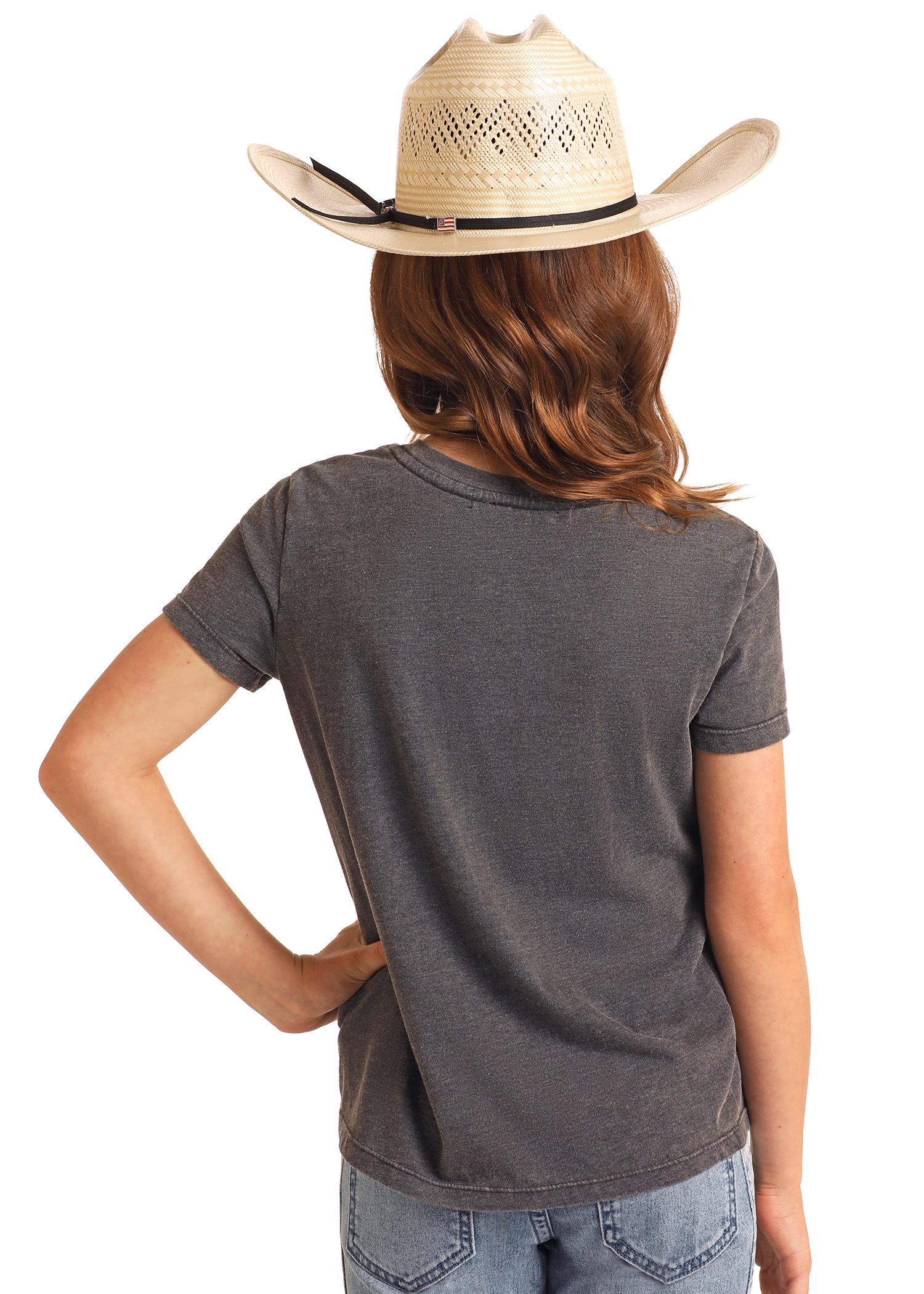 A person with wavy brown hair in a beige cowboy hat, blue jeans, and the Rock & Roll Denim Girls' Aztec Horse Graphic T-Shirt stands facing away with one hand on their hip against a plain background.