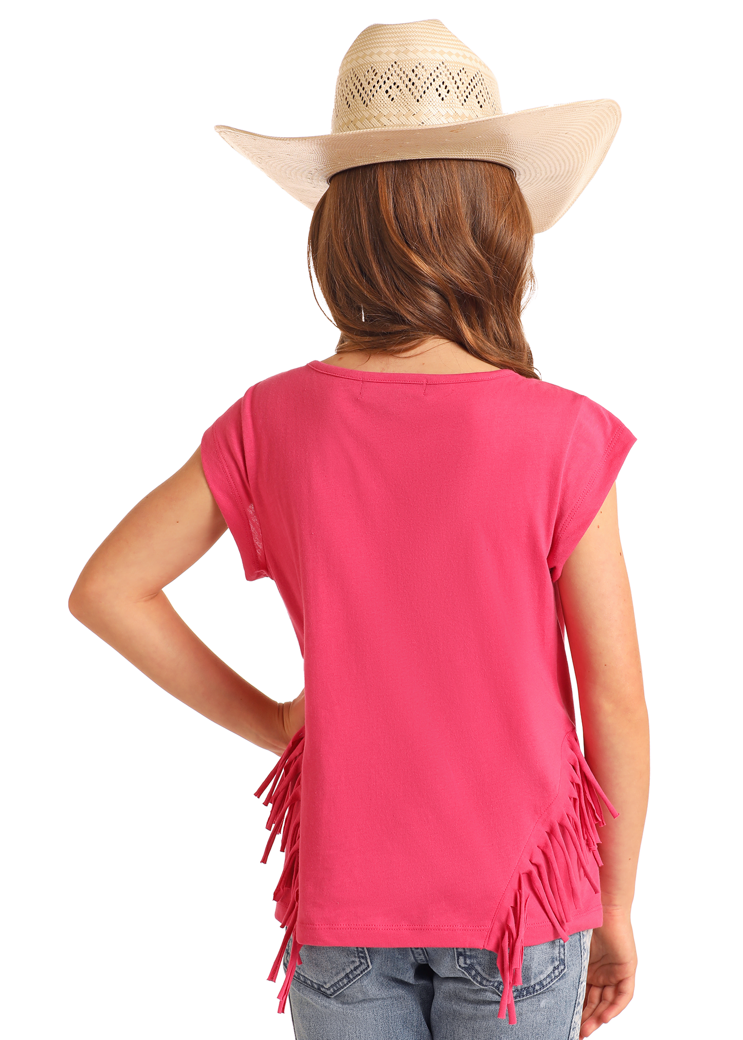 A young girl in a beige cowboy hat, blue jeans, and a Rock & Roll Denim Girls' Rhinestone Desert Graphic T-Shirt with bright pink fringe stands with her hand on her hip, facing away from the camera.