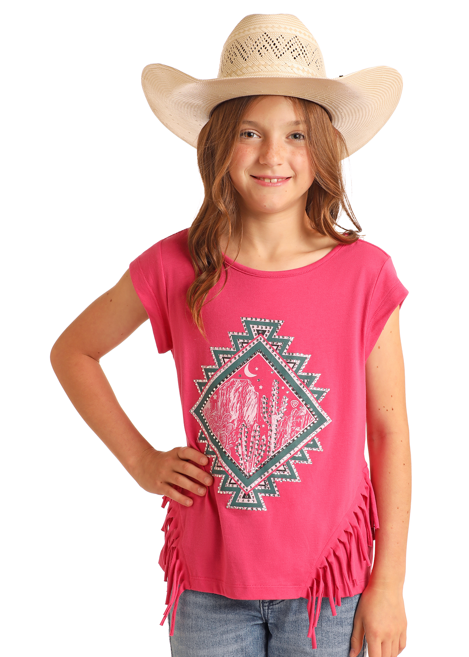 A smiling young girl in a wide-brimmed cowboy hat, blue jeans, and a Rock & Roll Denim Girls' Rhinestone Desert Graphic T-Shirt stands confidently with one hand on her hip against a plain background.