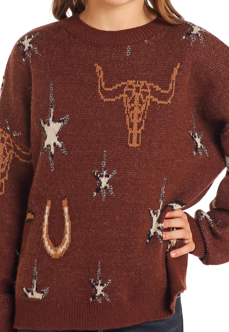 A girl models the Rock & Roll Denim Girls' Western Print Sweater, featuring bull skull, horseshoe, and star designs. Only her torso is visible, with one hand resting on her hip.