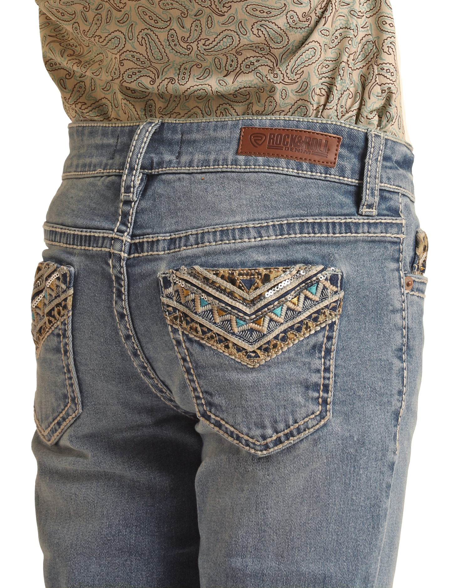A person wears Rock & Roll Denim Girls' Leopard Embroidered Pocket Bootcut Jeans with decorative stitching and beadwork on the back pockets, a brand leather patch on the waistband, paired with a patterned shirt tucked in.