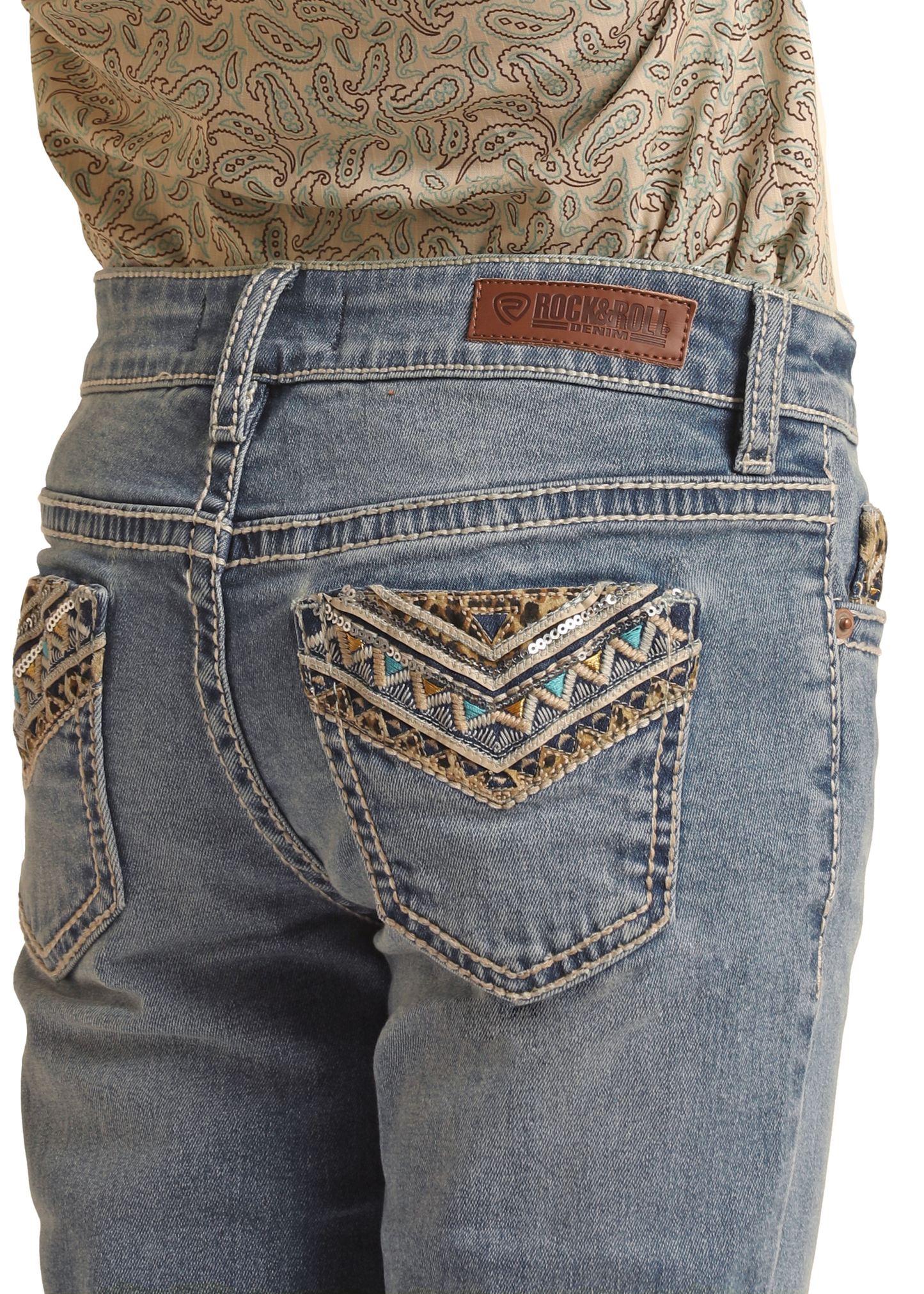 A person wears Rock & Roll Denim Girls' Leopard Embroidered Pocket Bootcut Jeans with decorative stitching and beadwork on the back pockets, a brand leather patch on the waistband, paired with a patterned shirt tucked in.