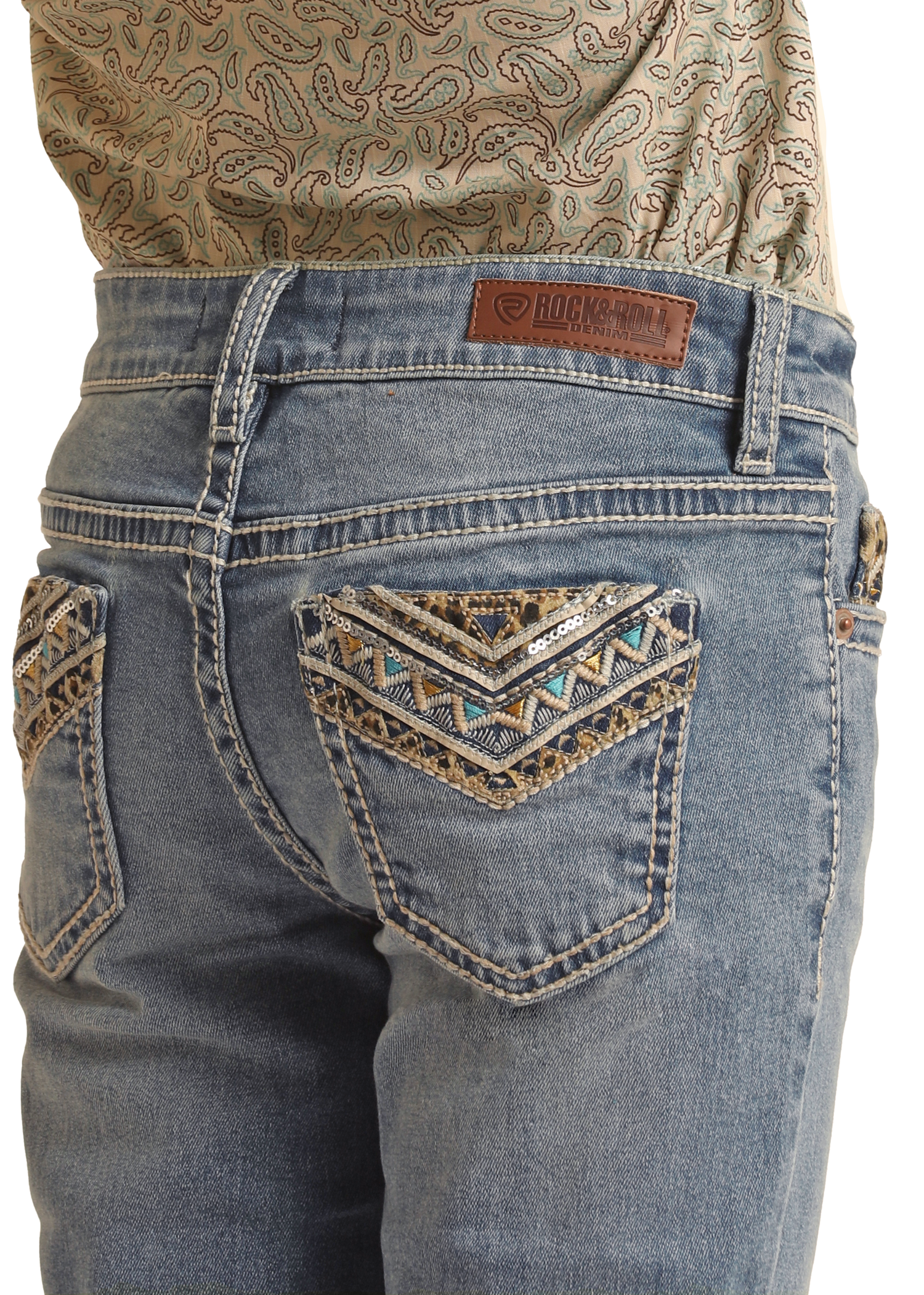 A person wears Rock & Roll Denim Girls' Leopard Embroidered Pocket Bootcut Jeans with decorative stitching and beadwork on the back pockets, a brand leather patch on the waistband, paired with a patterned shirt tucked in.