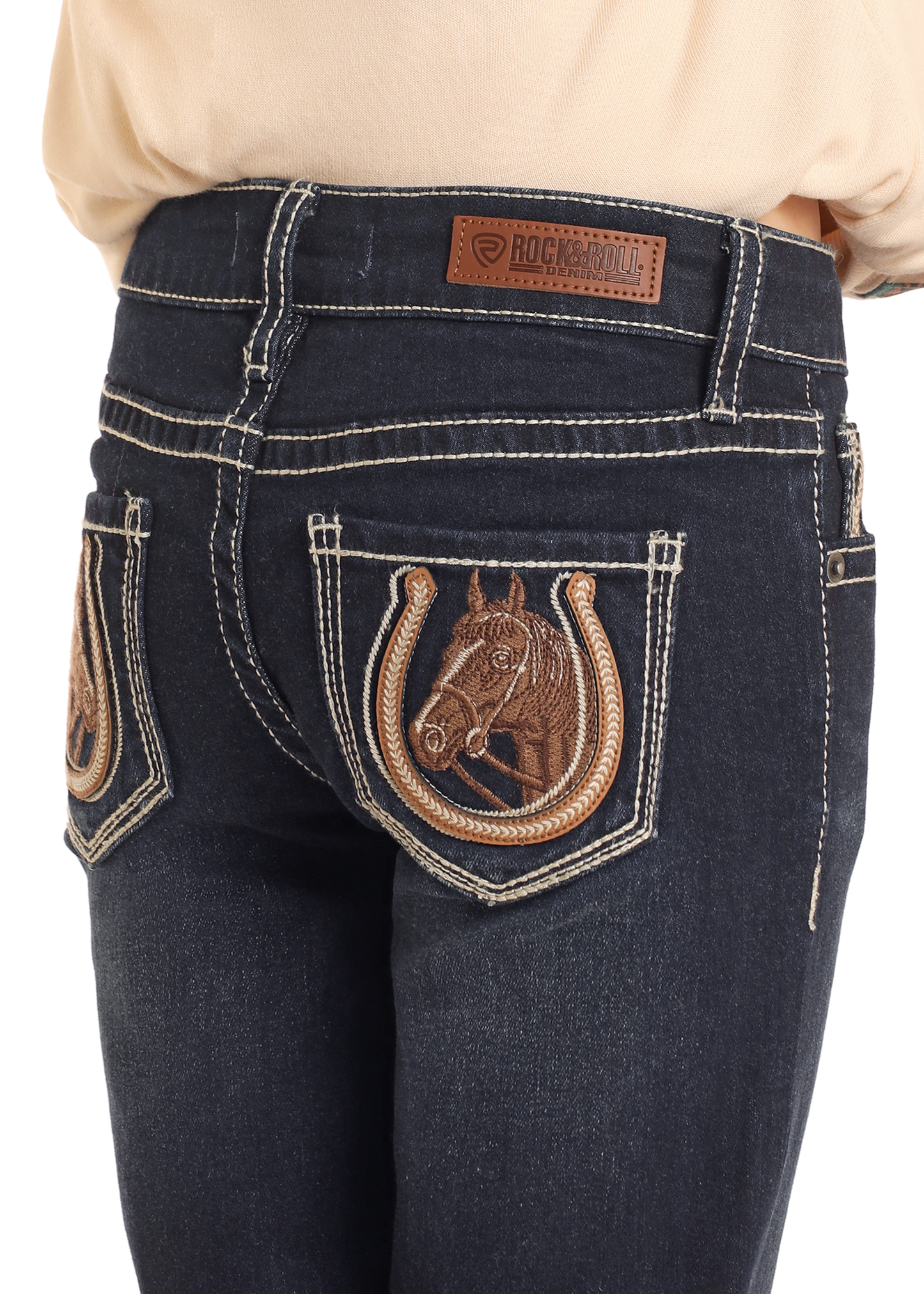 A close-up shows a girl wearing Rock & Roll Denim Girls' Mid Rise Extra Stretch Horse Bootcut Jeans, featuring white stitching, back pockets with a horse head in a horseshoe, and a brown leather brand patch on the waistband.