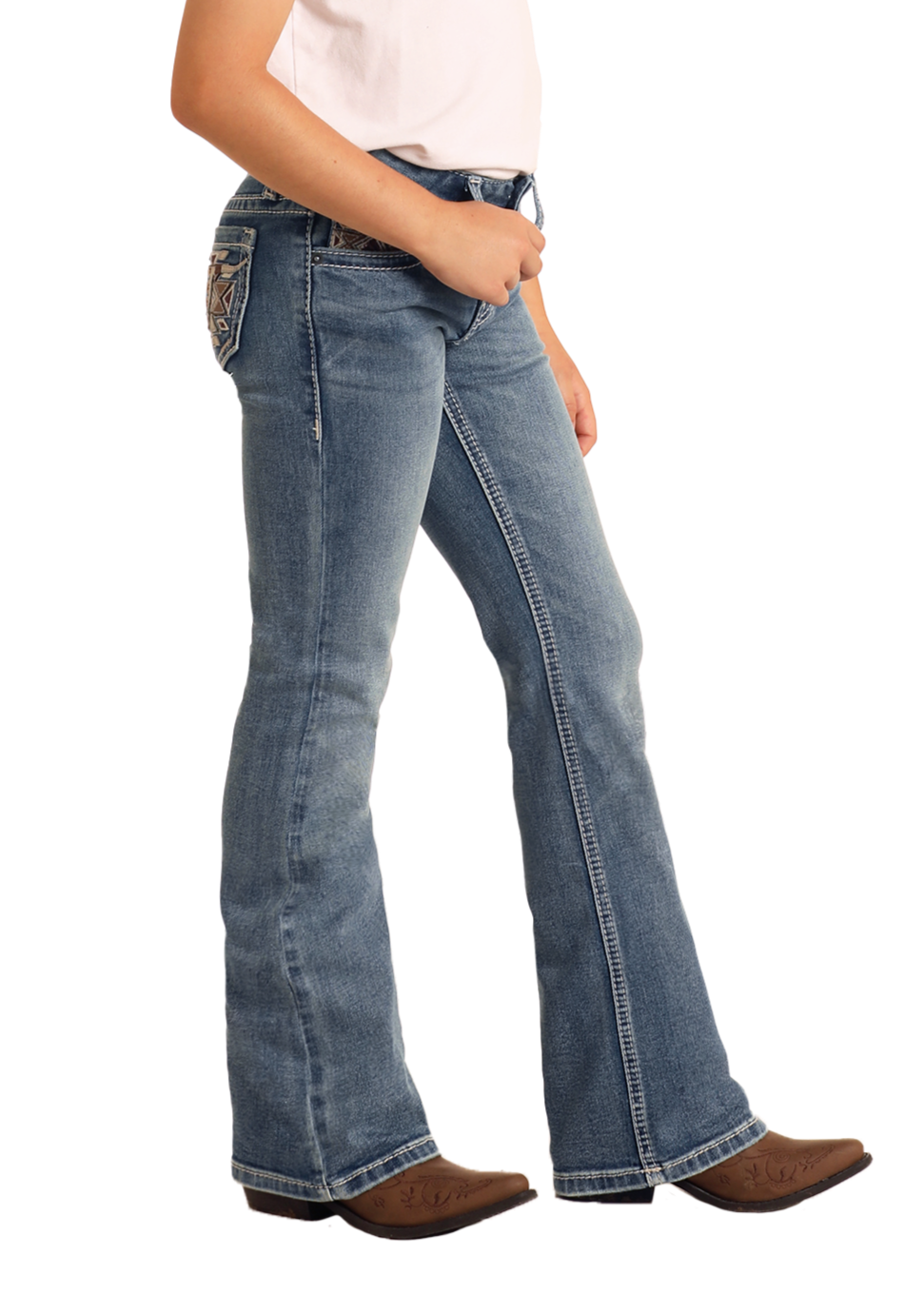 A girl models Rock & Roll Denim Girls' Mid Rise Steer Head Embroidered Bootcut Jeans with decorative back stitching, brown cowboy boots, and a light-colored shirt against a plain backdrop; only her lower half is shown.