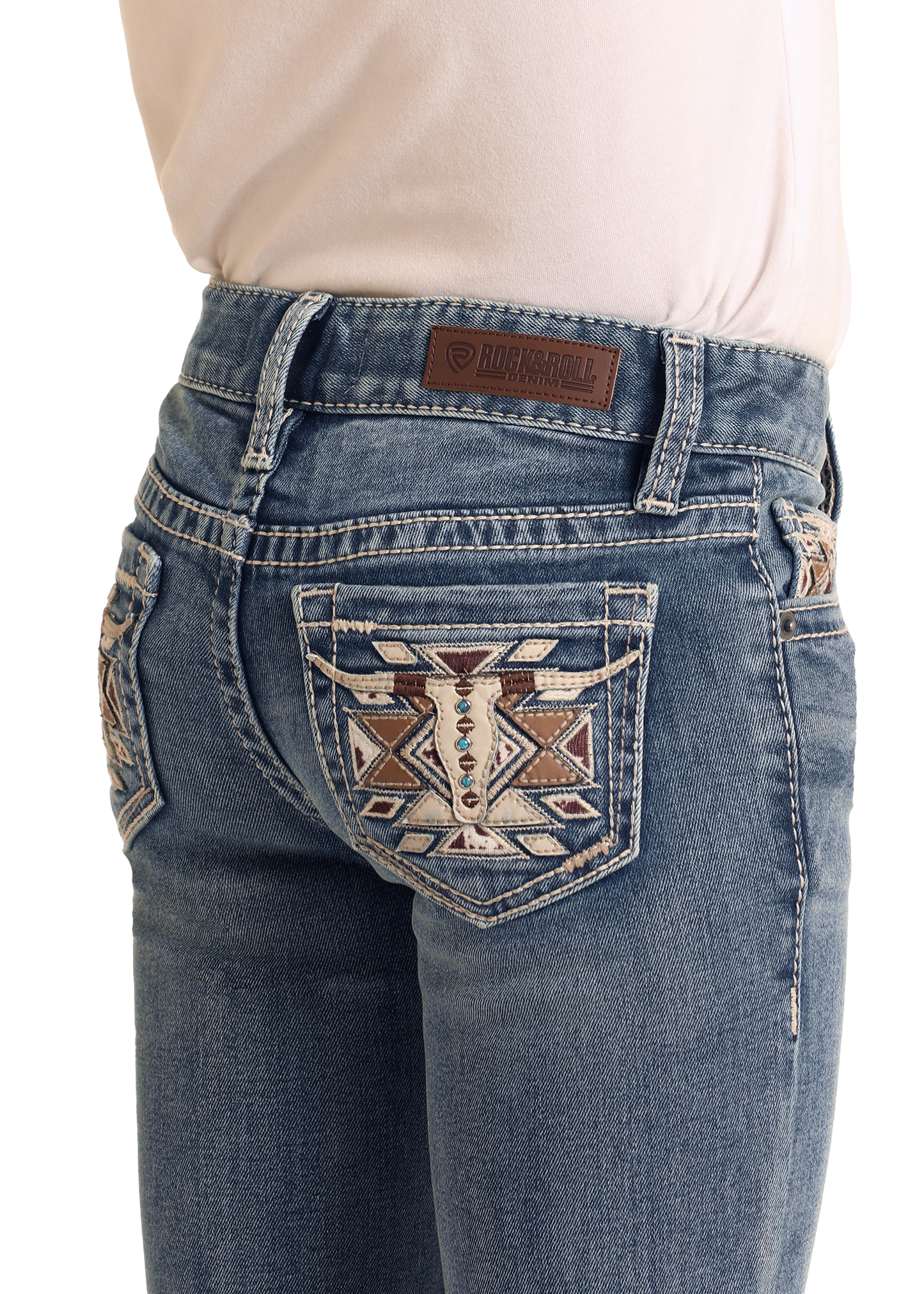 A girl wears Rock & Roll Denim Girls' Mid Rise Steer Head Embroidered Bootcut Jeans with decorative back pocket stitching and a brown leather brand patch, paired with a light pink tucked-in shirt for a classic look.