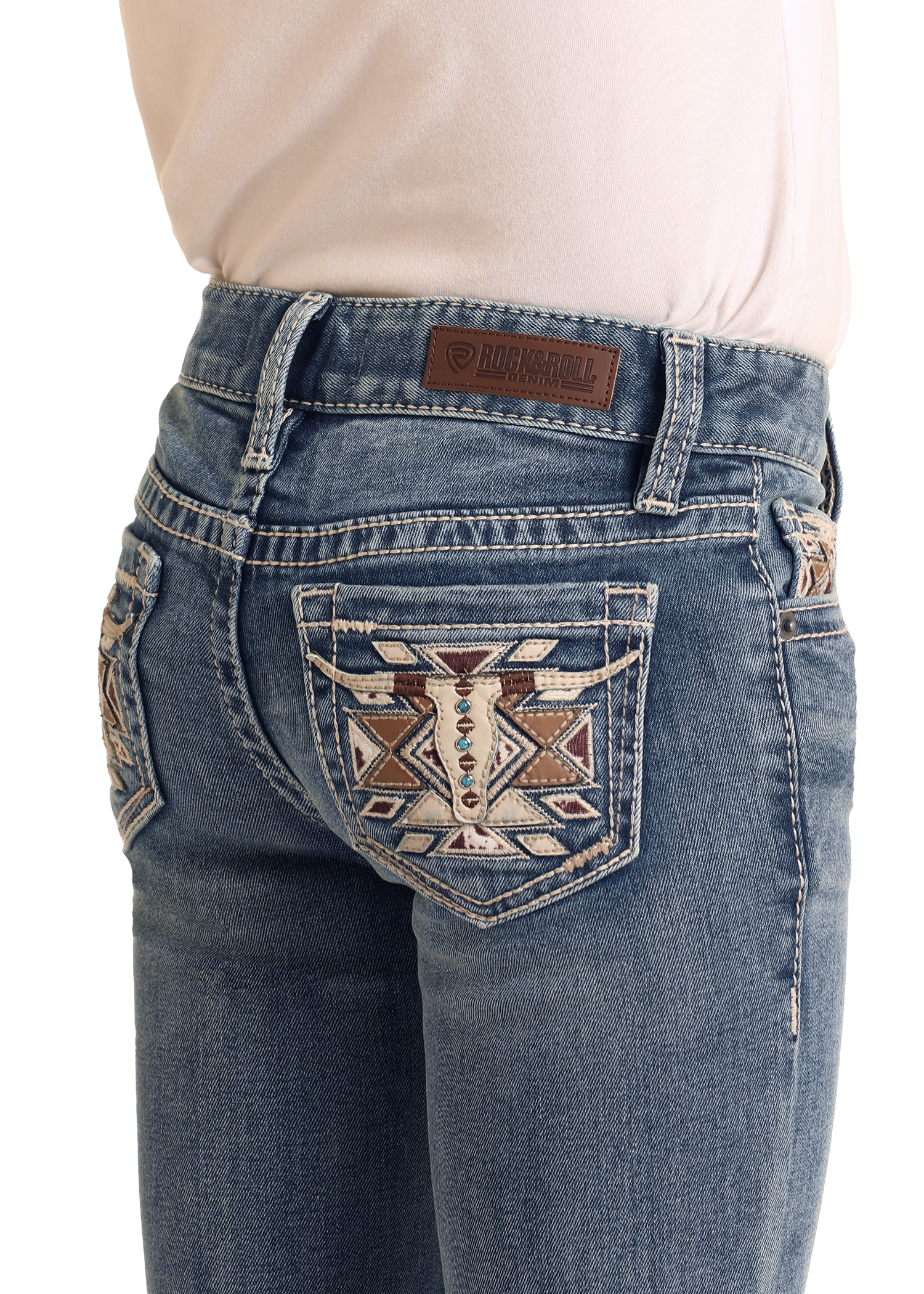 A girl wears Rock & Roll Denim Girls' Mid Rise Steer Head Embroidered Bootcut Jeans with decorative back pocket stitching and a brown leather brand patch, paired with a light pink tucked-in shirt for a classic look.