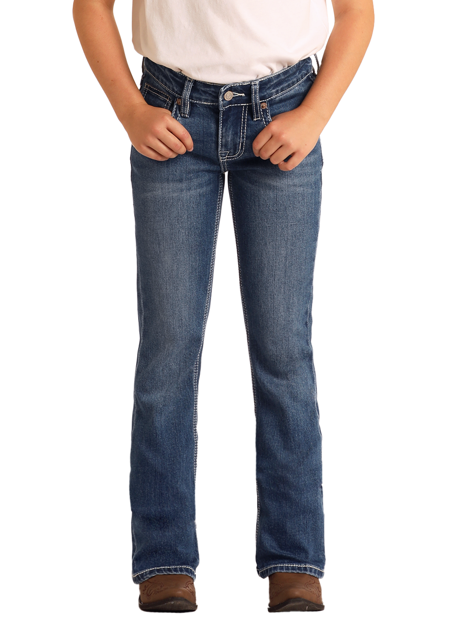 A girl wearing Rock & Roll Denim Girls' Embroidered Denim Star Bootcut Jeans stands against a white background, hands near her pockets, paired with a white shirt and brown shoes.