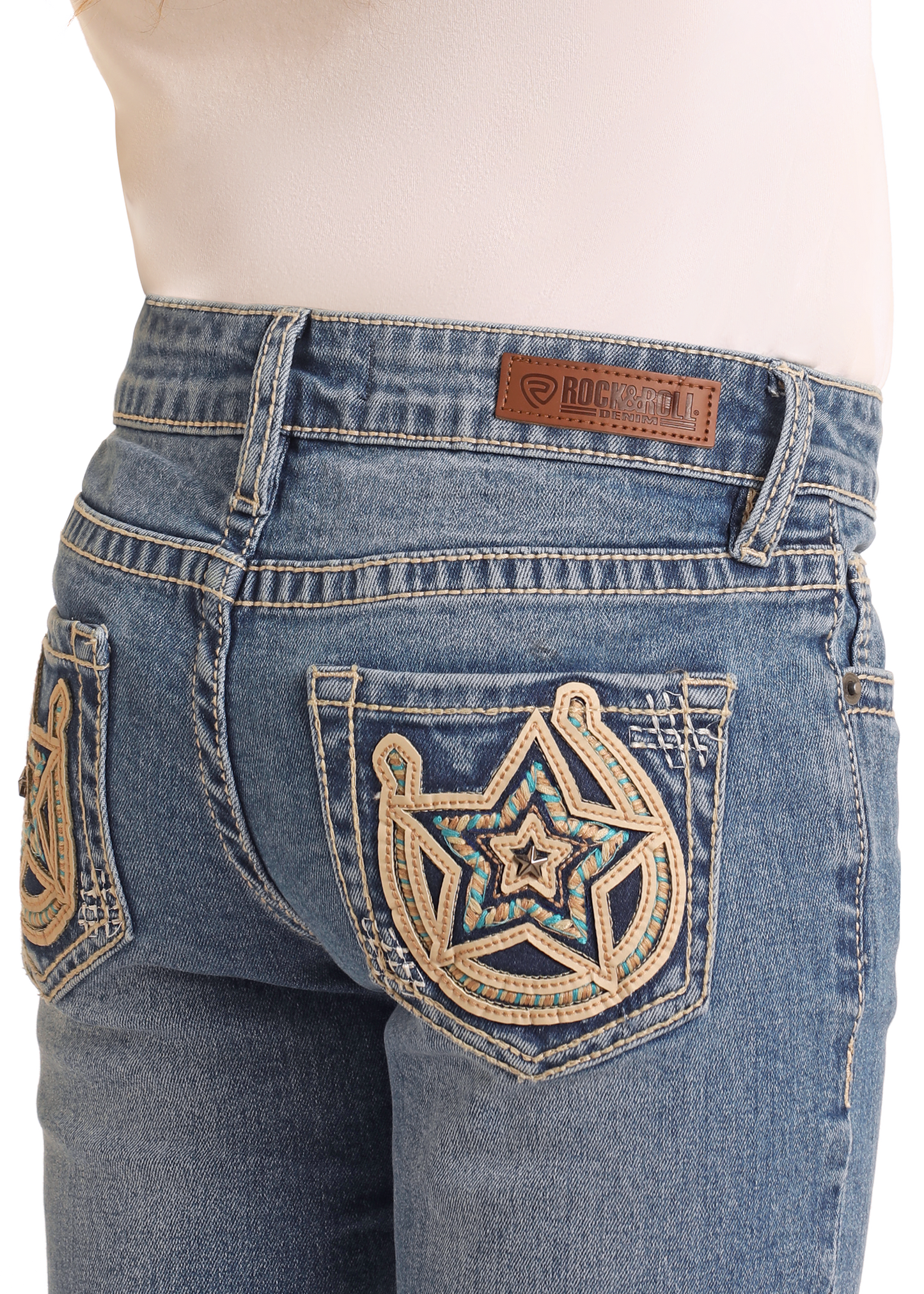 A person wears Rock & Roll Denim Girls' Mid Rise Extra Stretch Horseshoe Star Bootcut Jeans in a medium wash, featuring Western-inspired star stitching, turquoise beads on the back pockets, and a brown leather brand label above the right pocket.