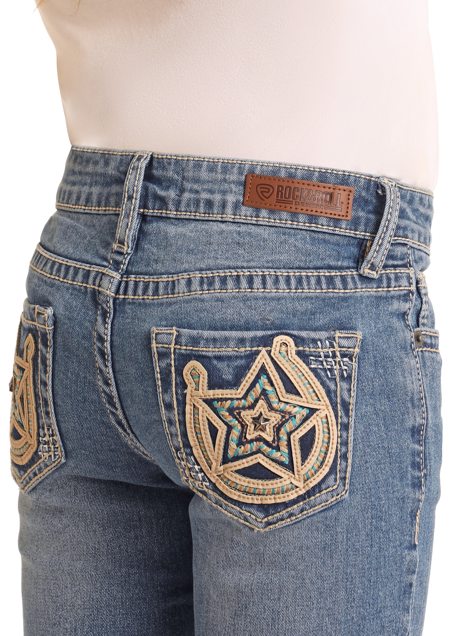 A person wears Rock & Roll Denim Girls' Mid Rise Extra Stretch Horseshoe Star Bootcut Jeans in a medium wash, featuring Western-inspired star stitching, turquoise beads on the back pockets, and a brown leather brand label above the right pocket.