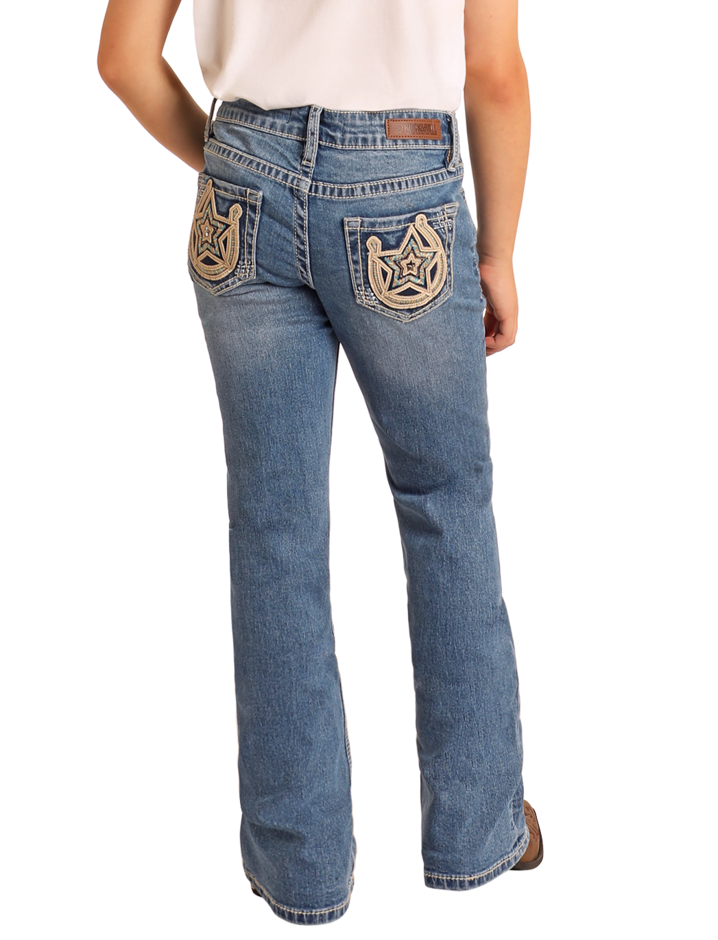 A person wearing Rock & Roll Denim Girls' Mid Rise Extra Stretch Horseshoe Star Bootcut Jeans with medium wash and white embroidered back pockets, paired with a white shirt, stands facing away.