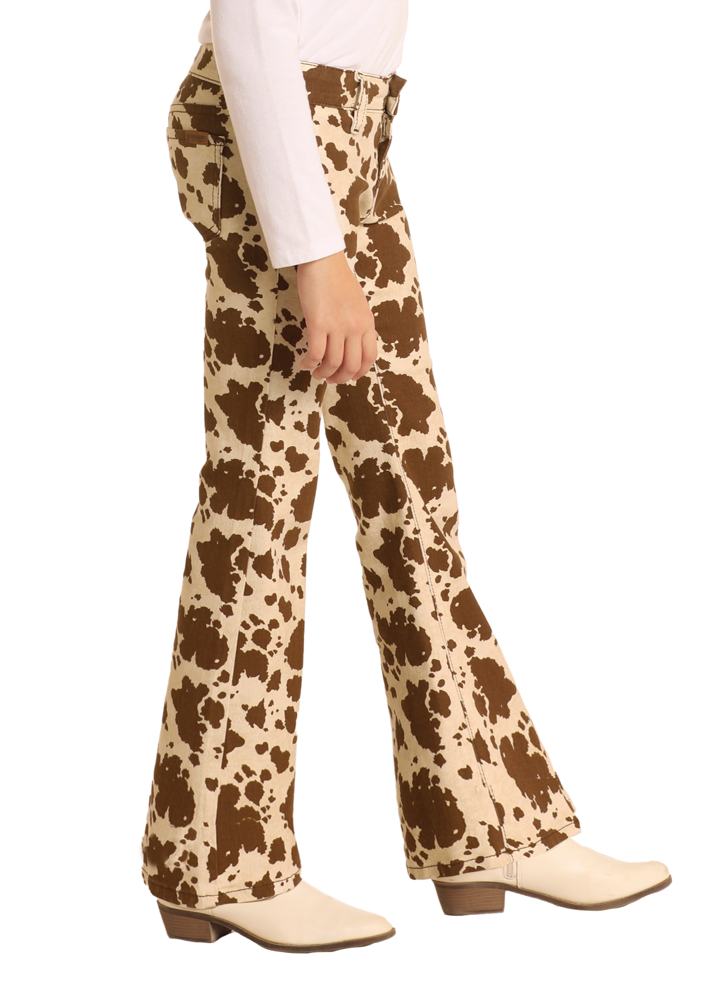 A person wears Rock & Roll Denim Girls' Cowhide Print Flare Jeans with a white long-sleeve top and cream heeled boots, standing against a plain white background.