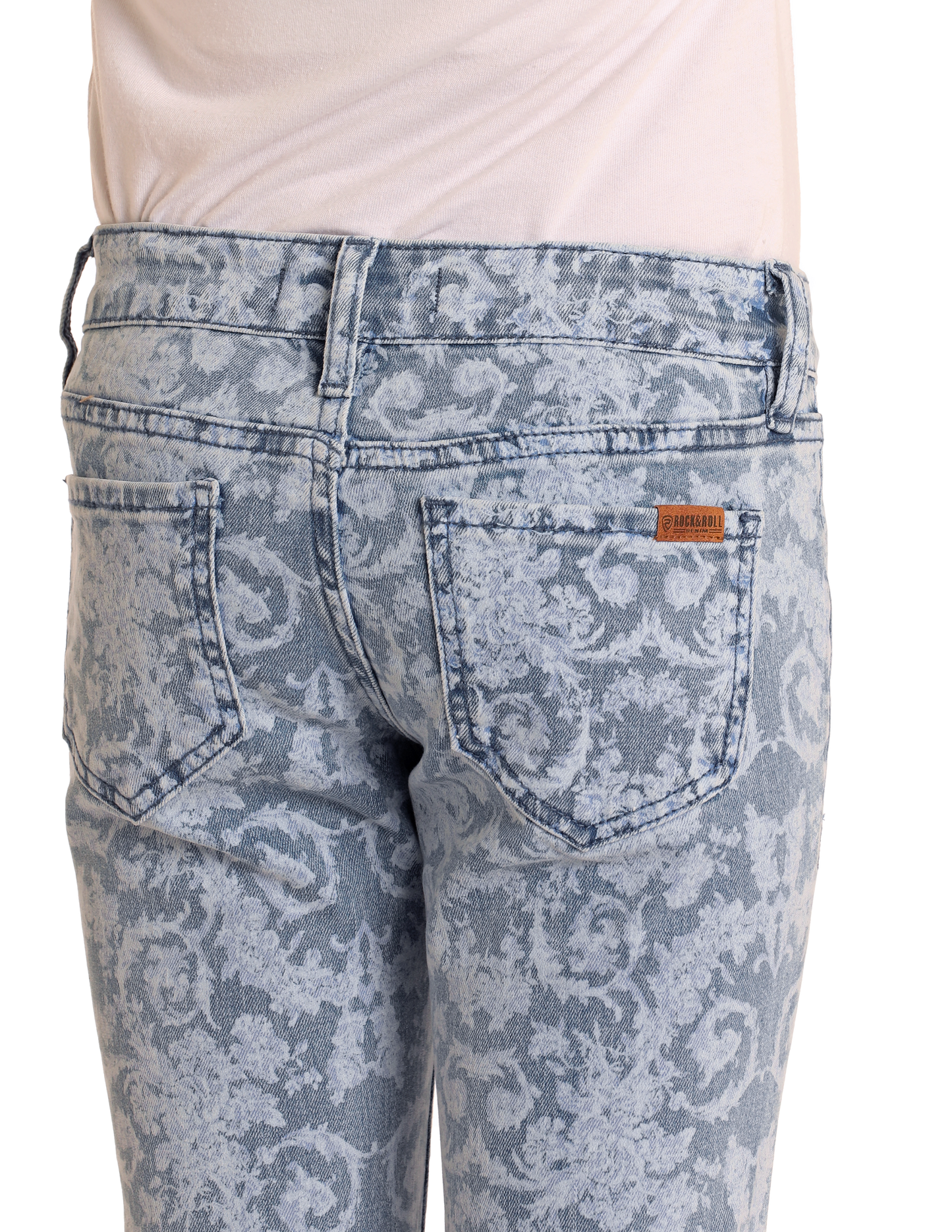 A close-up of the back of a person wearing Rock & Roll Denim Girls' Floral Print Flare Jeans in medium wash, featuring two pockets, a subtle floral print, and a tan brand label above the right pocket. The top is neatly tucked in for style.