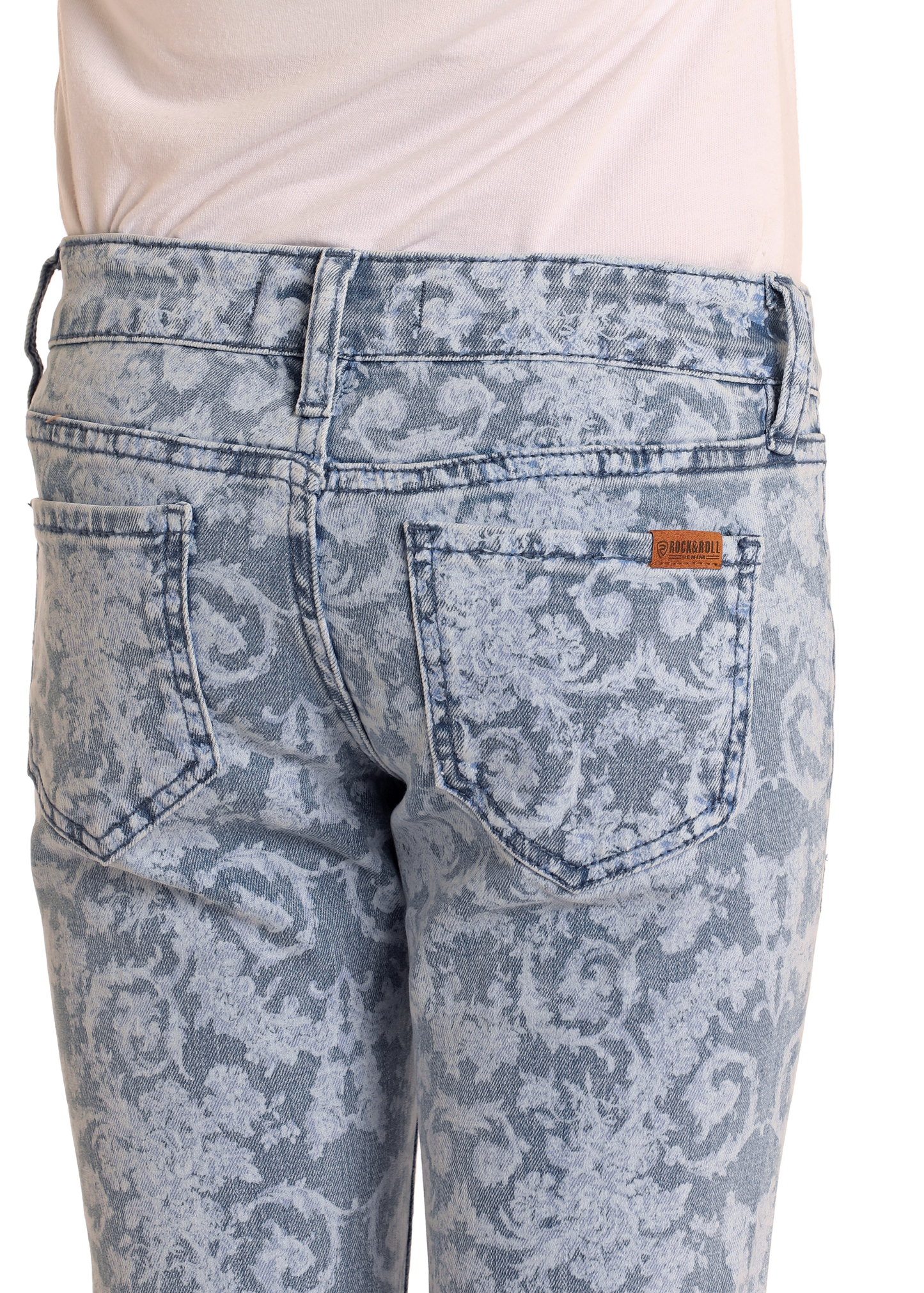 A close-up of the back of a person wearing Rock & Roll Denim Girls' Floral Print Flare Jeans in medium wash, featuring two pockets, a subtle floral print, and a tan brand label above the right pocket. The top is neatly tucked in for style.