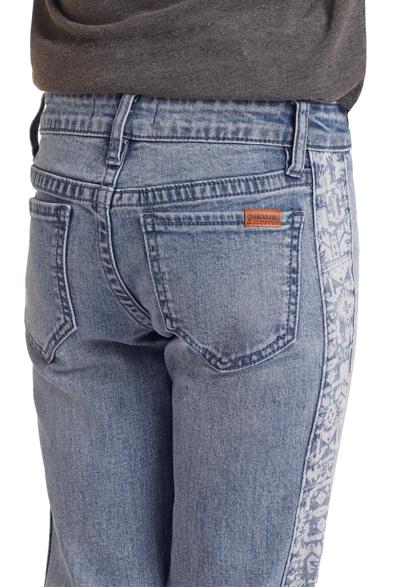 Close-up of a person wearing Rock & Roll Denim Girls' Aztec Panel Button Flare Jeans in light blue, featuring a faded patterned side panel and a small brown label above the right pocket. A tucked-in dark shirt completes the Southwestern look.