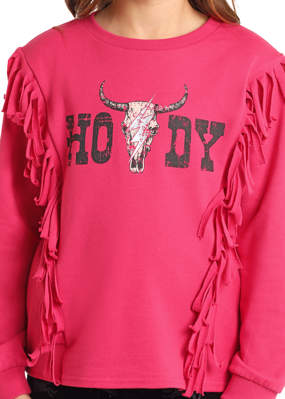 A person wears a bright pink Rock & Roll Denim Girls' Howdy Fringe Graphic Pullover, featuring fringe sleeves, a bull skull graphic, and "HOWDY" in distressed black letters across the chest.