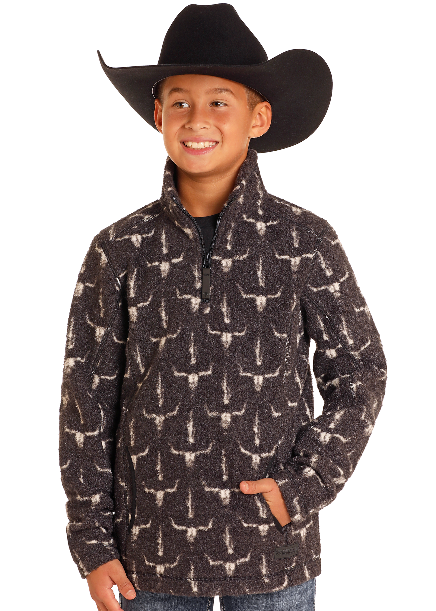 A smiling child in a large black cowboy hat and the Rock & Roll Denim Kids' Steer Skull Quarter Zip Pullover, hands in pockets, stands against a plain white background.