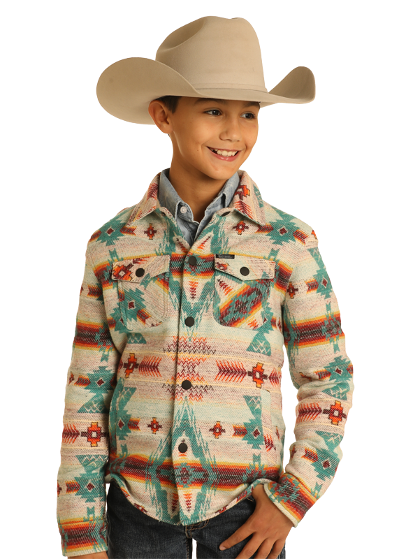 A smiling child wears the Rock & Roll Denim Kids' Cream Aztec Jacquard Shirt Jacket, featuring colorful southwestern designs, a beige cowboy hat, and denim shirt, standing with one hand in their pocket against a plain background.