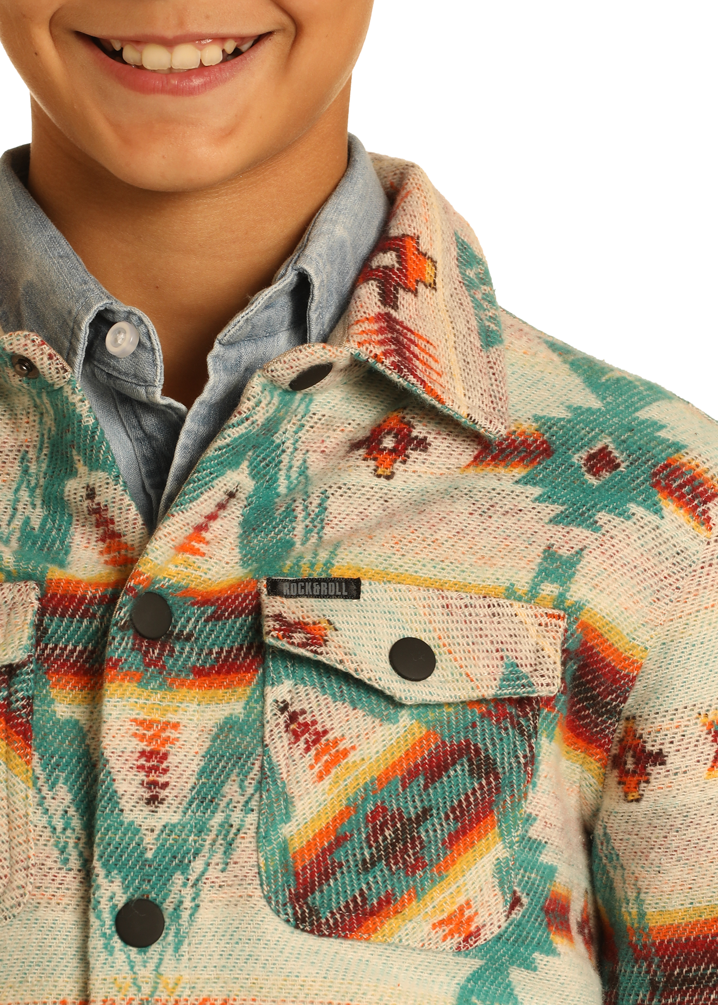 A close-up of a smiling child in the Rock & Roll Denim Kids' Cream Aztec Jacquard Shirt Jacket, featuring turquoise, orange, and red designs over a blue button-up, with focus on the 100% cotton jacket’s front pocket and collar.